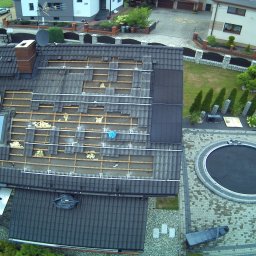 Krukterm Sp. z. o. o - Widok z góry na dach domu w trakcie prac remontowych, częściowo zdjęte dachówki, widoczne fragmenty ocieplenia, pracownik na podeście, ogród z basenem w tle.