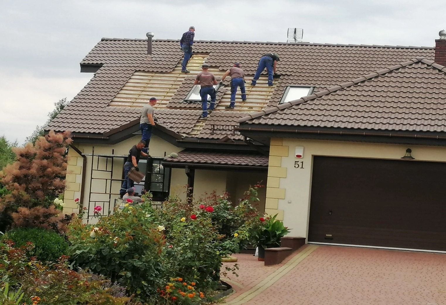 Ekipa dekarzy wymienia dachówkę na brązowym dachu domu jednorodzinnego z oknami dachowymi; widok z poziomu ogrodu z kwitnącymi różami i innymi roślinami.