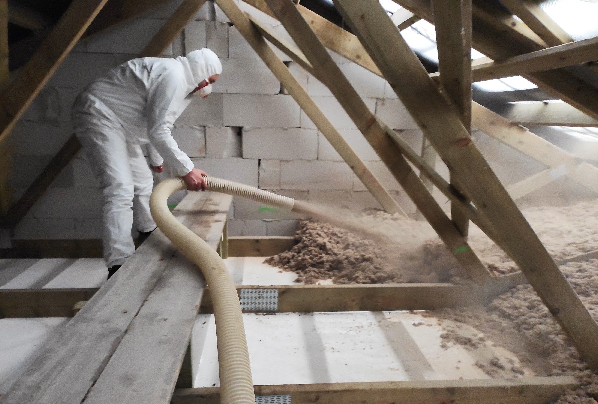 Pracownik w kombinezonie ochronnym aplikuje materiał izolacyjny za pomocą węża na poddaszu z widoczną konstrukcją drewnianą i bloczkami z betonu komórkowego w tle.