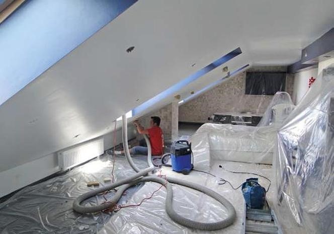 Pracownik w czerwonej koszulce aplikuje izolację natryskową na skośny sufit poddasza, widoczny agregat natryskowy i zabezpieczone folią meble.