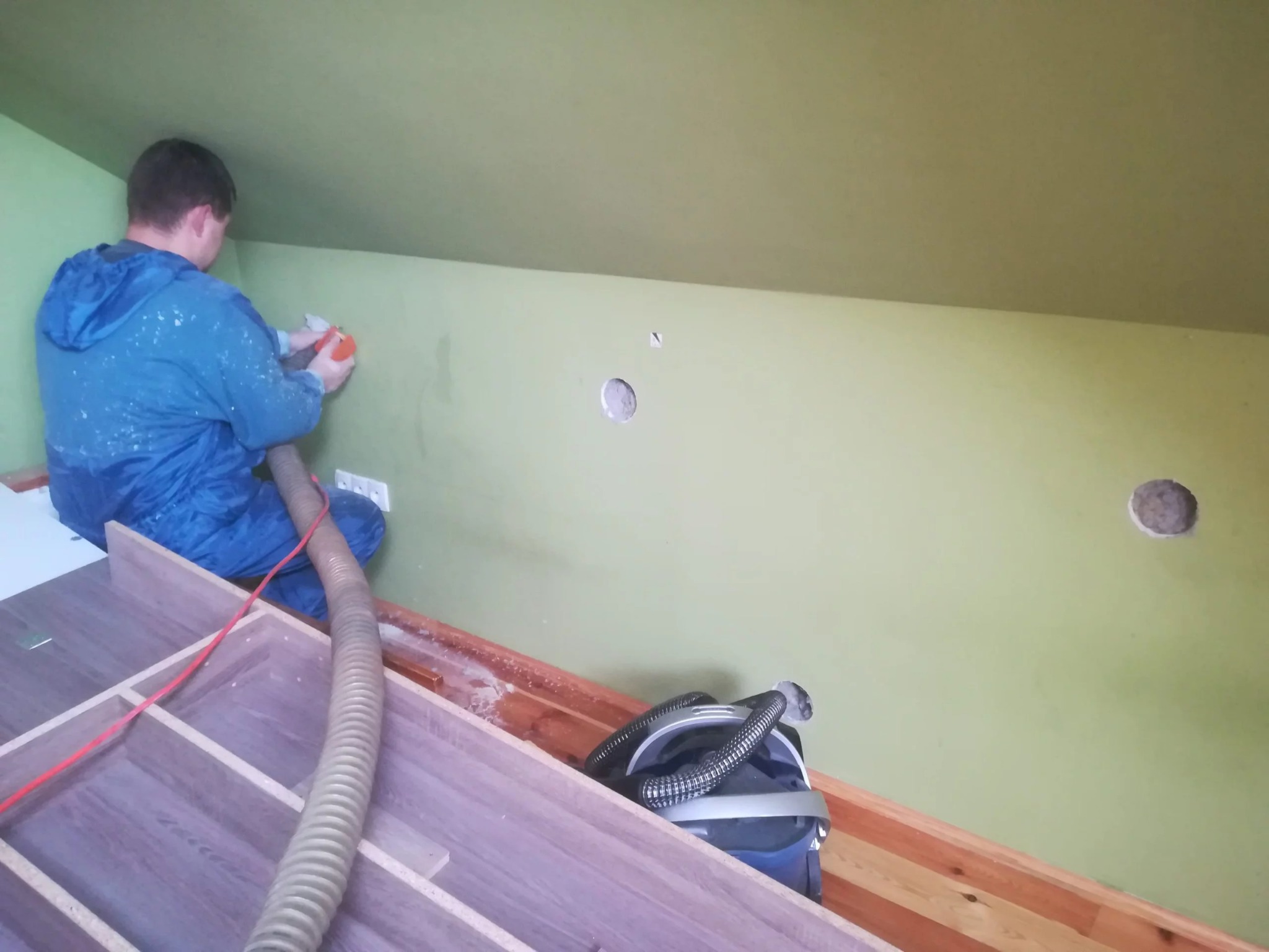 Pracownik w niebieskim kombinezonie aplikuje materiał izolacyjny w otwór w zielonej ścianie na poddaszu, widoczny odkurzacz przemysłowy i elementy konstrukcyjne mebli.