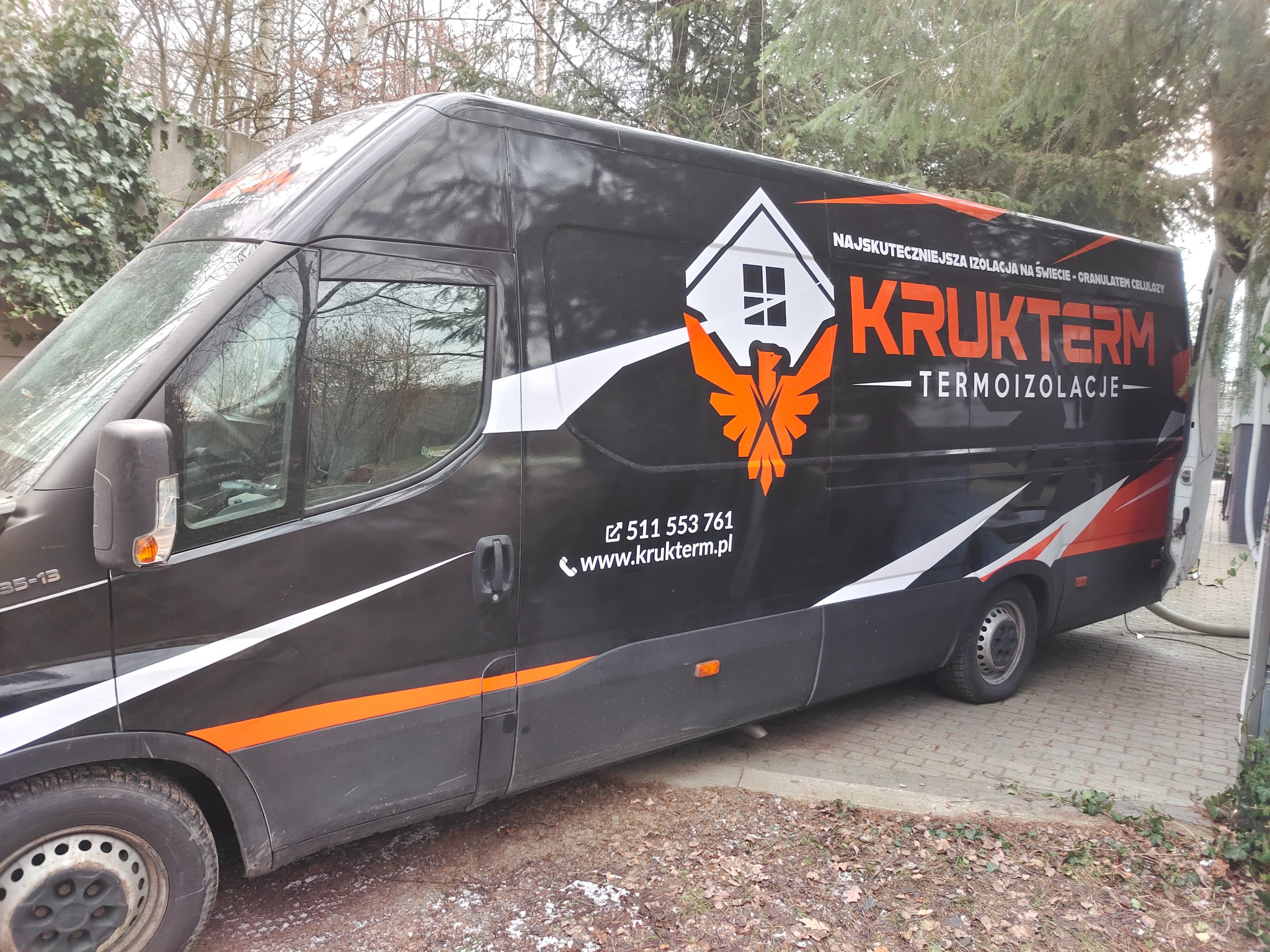 Czarny van firmy Krukterm z logo i numerem telefonu, zaparkowany na brukowanej powierzchni, z widocznym wężem do wdmuchiwania izolacji.