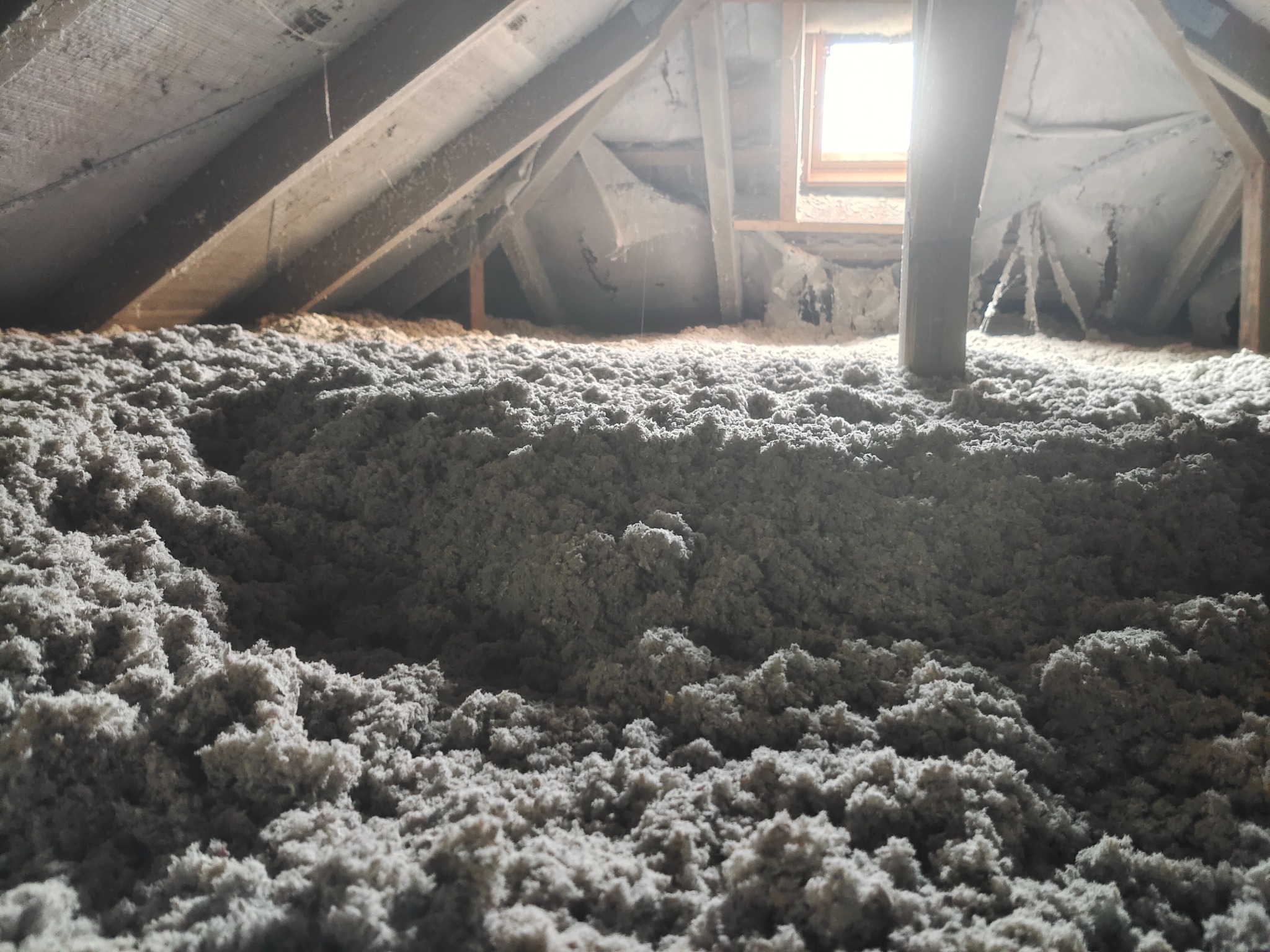 Szara, włóknista izolacja termiczna wypełniająca przestrzeń poddasza z widocznymi elementami drewnianej konstrukcji dachu i małym oknem w tle.