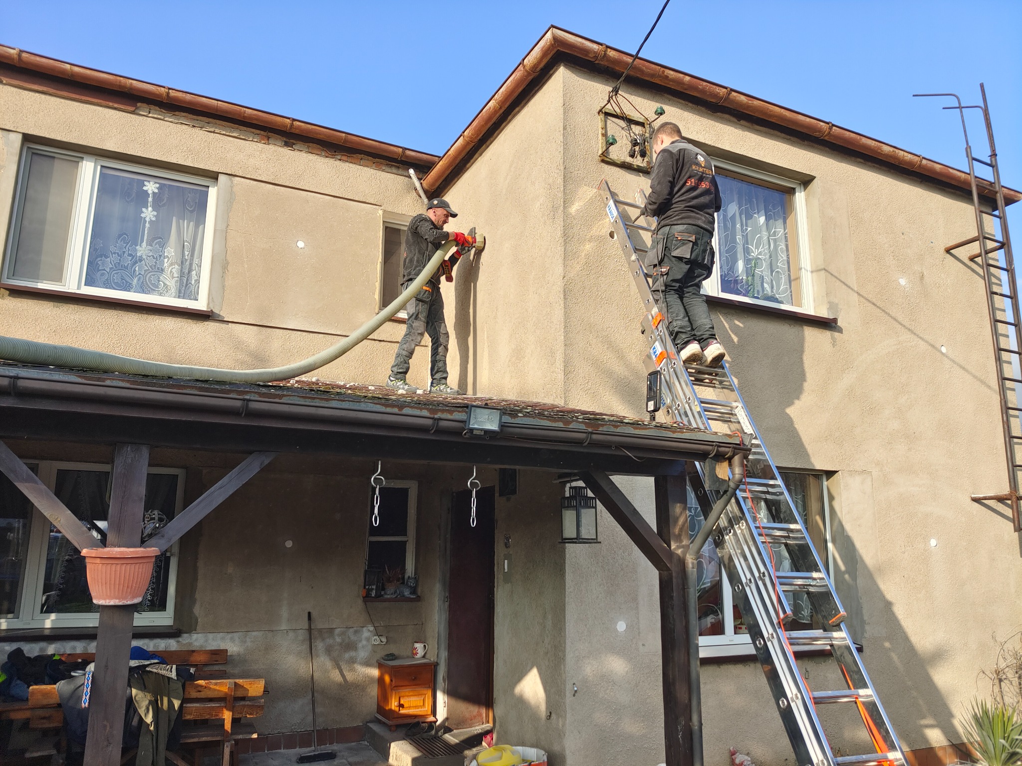 Ocieplanie ściany budynku pianką PUR przez dwóch pracowników w kombinezonach, jeden aplikuje pianę z węża, drugi stoi na drabinie aluminiowej, widoczne okno z firanką i stary budynek z szarą elewacją.