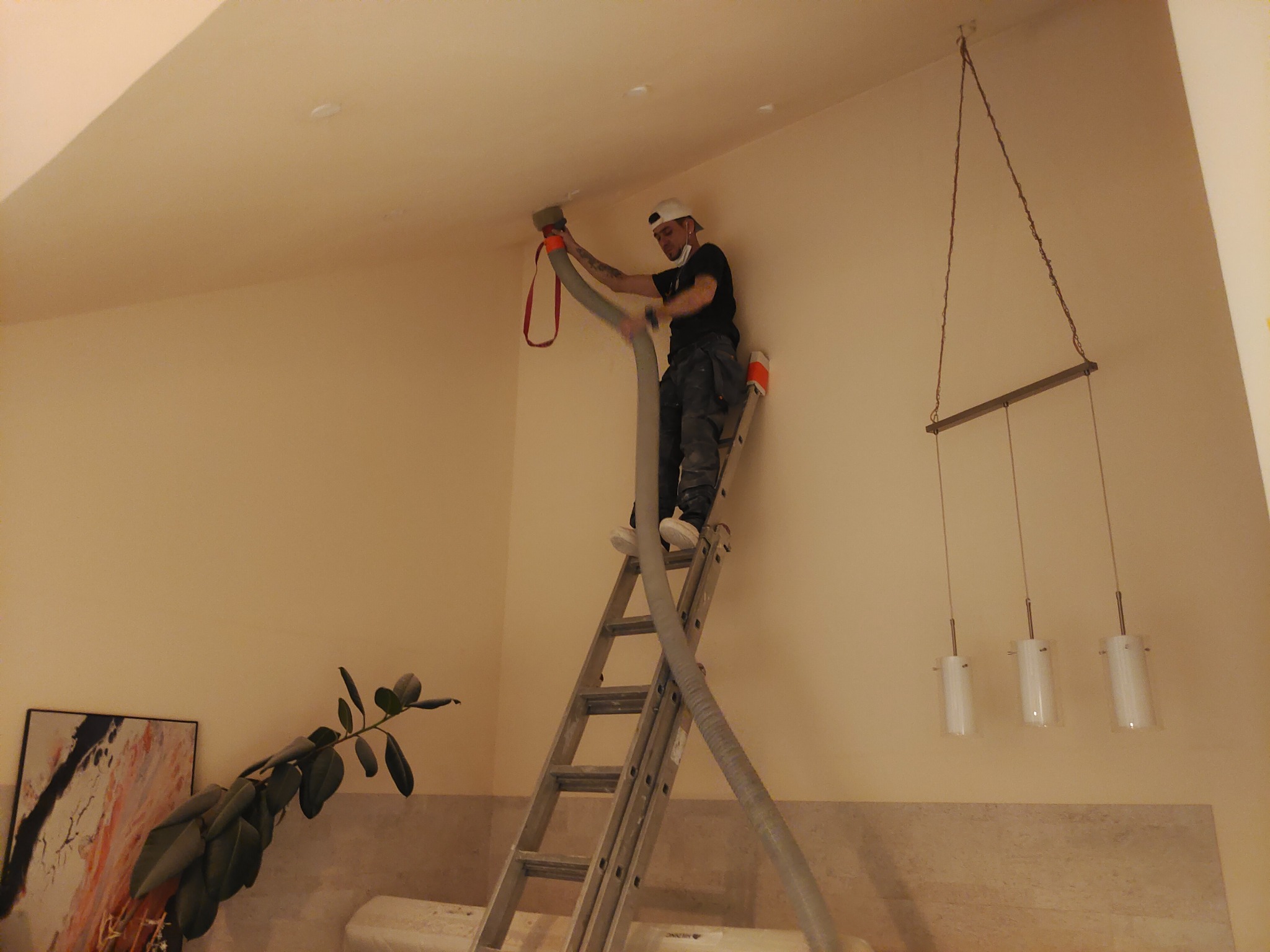 Pracownik na drabinie aplikuje materiał izolacyjny na suficie za pomocą węża, widoczny element oświetlenia i roślina w tle.