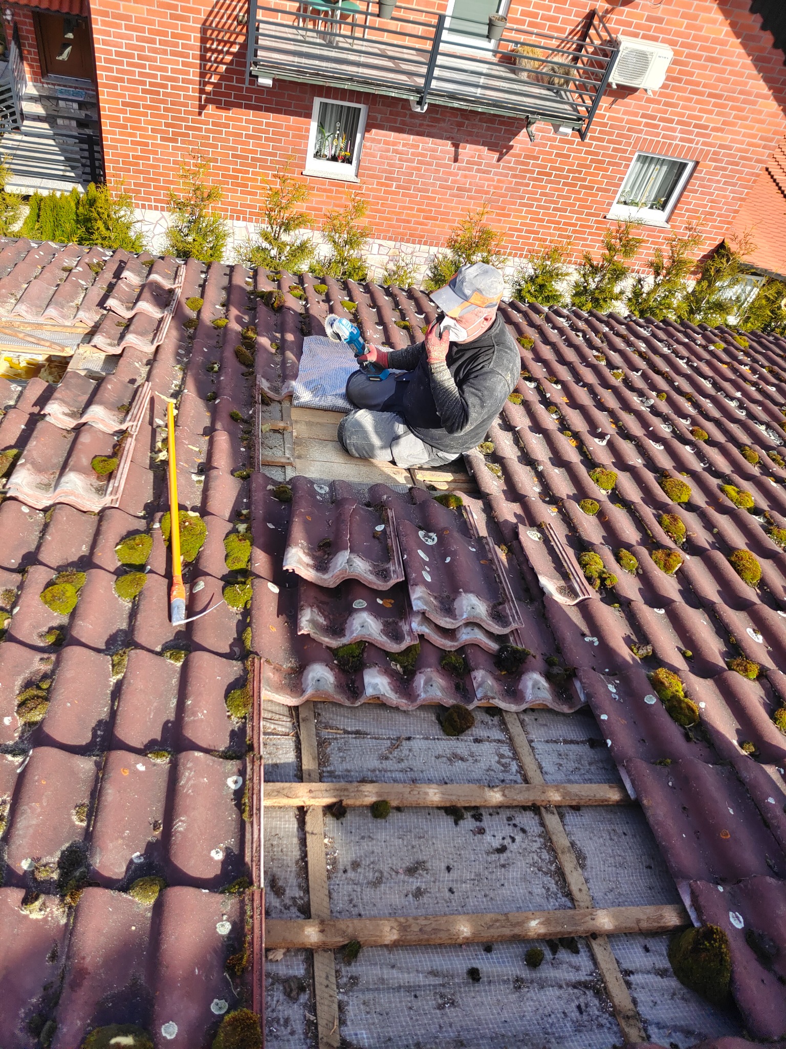 Pracownik w masce i czapce siedzi na dachu z czerwonej dachówki pokrytej mchem, obok leży żółta miarka, a on trzyma w ręku szlifierkę kątową, przygotowując się do pracy nad odsłoniętą konstrukcją...