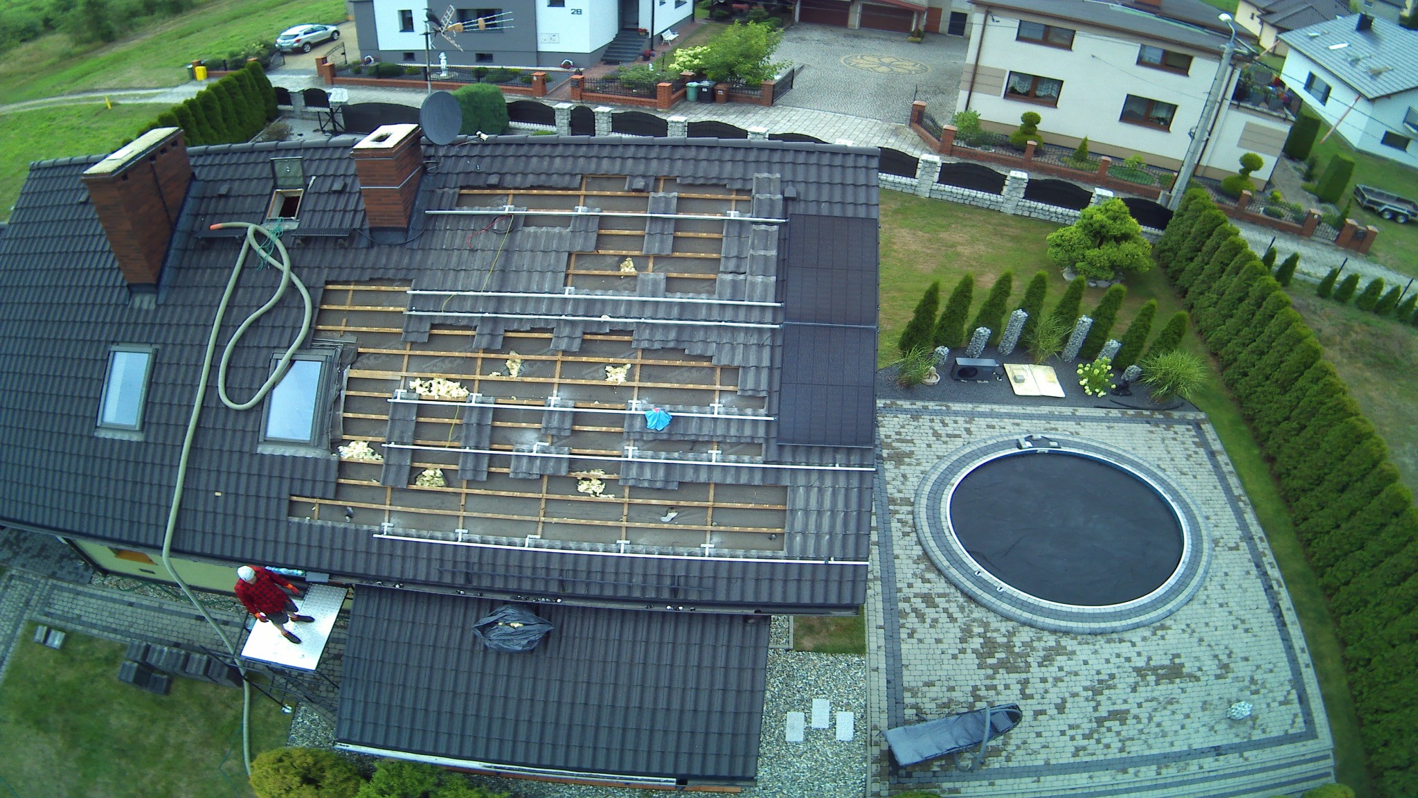 Widok z góry na dach domu w trakcie prac remontowych, częściowo zdjęte dachówki, widoczne fragmenty ocieplenia, pracownik na podeście, ogród z basenem w tle.
