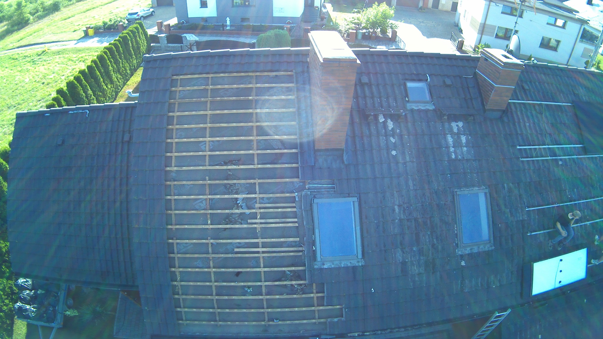 Widok z góry na remontowany dach z odsłoniętą konstrukcją drewnianą, widoczne okna dachowe, komin oraz pracownik na dachu.