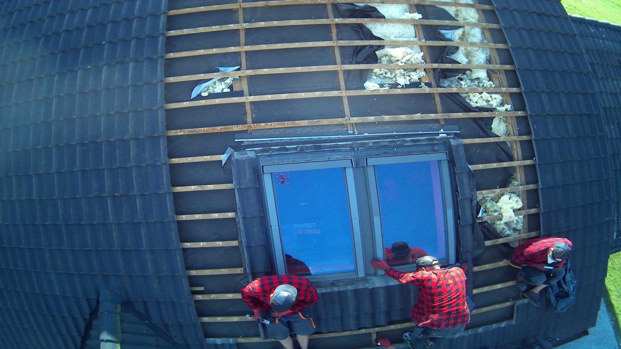 Montaż okna dachowego z widoczną izolacją termiczną pod konstrukcją dachu, trzech pracowników w czerwonych koszulach w kratę podczas pracy.