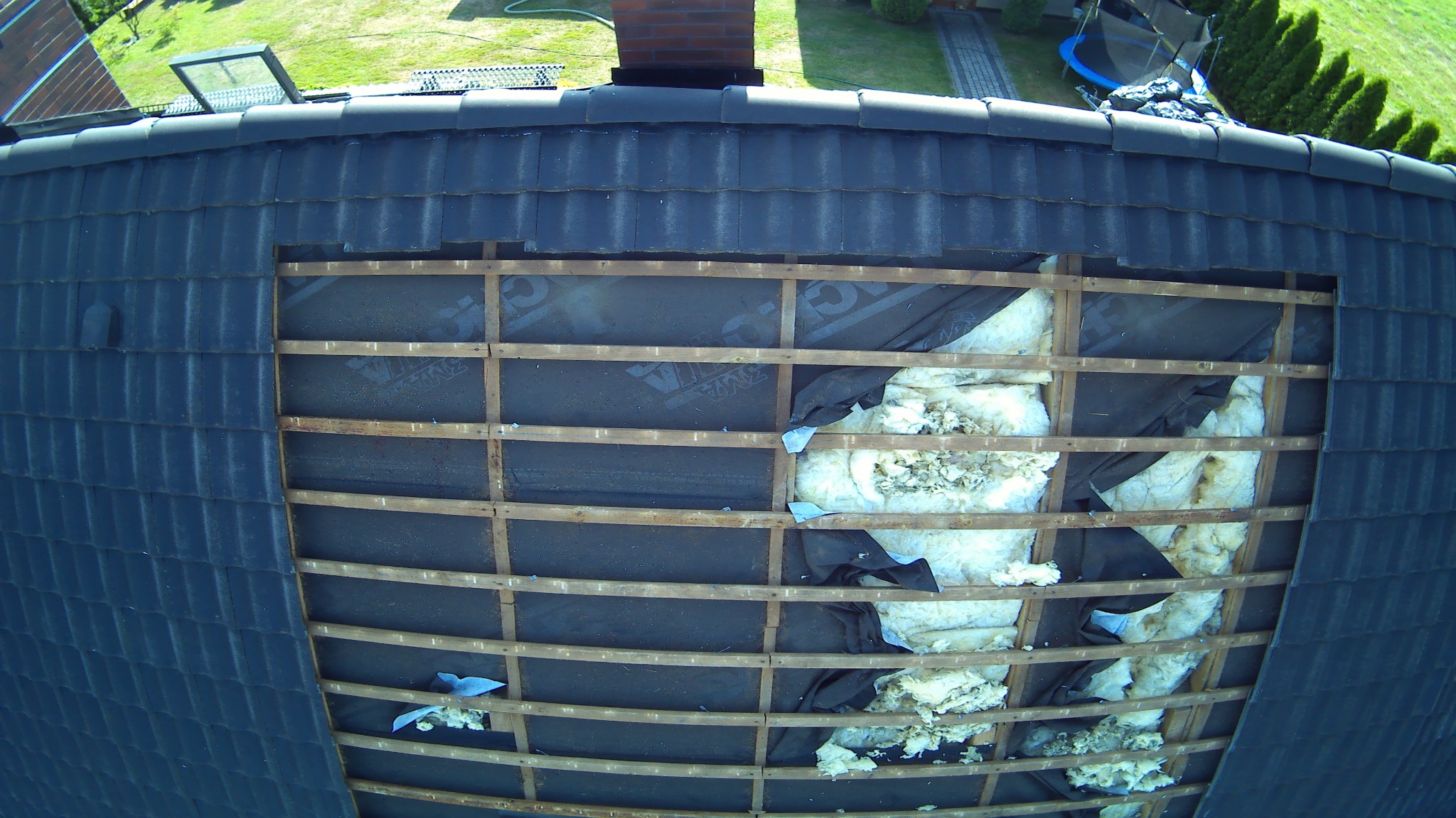 Fragment dachu z odsłoniętą konstrukcją i częściowo widoczną izolacją z wełny mineralnej, czarna membrana wiatroizolacyjna, widok z góry na tle trawnika i drzew