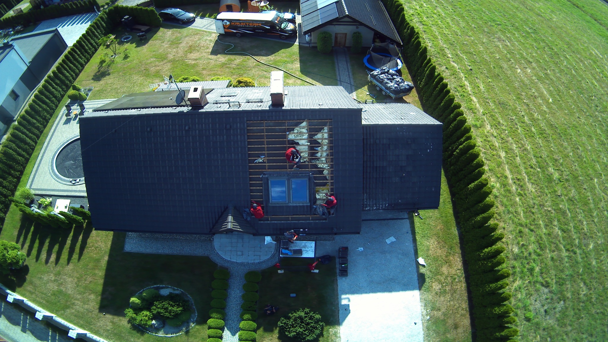 Widok z góry na ekipę w czerwonych kombinezonach ocieplającą poddasze pianką PUR, widoczna otwarta konstrukcja dachu, częściowo wypełniona izolacją, z boku zaparkowany samochód firmy ociepleniowej.