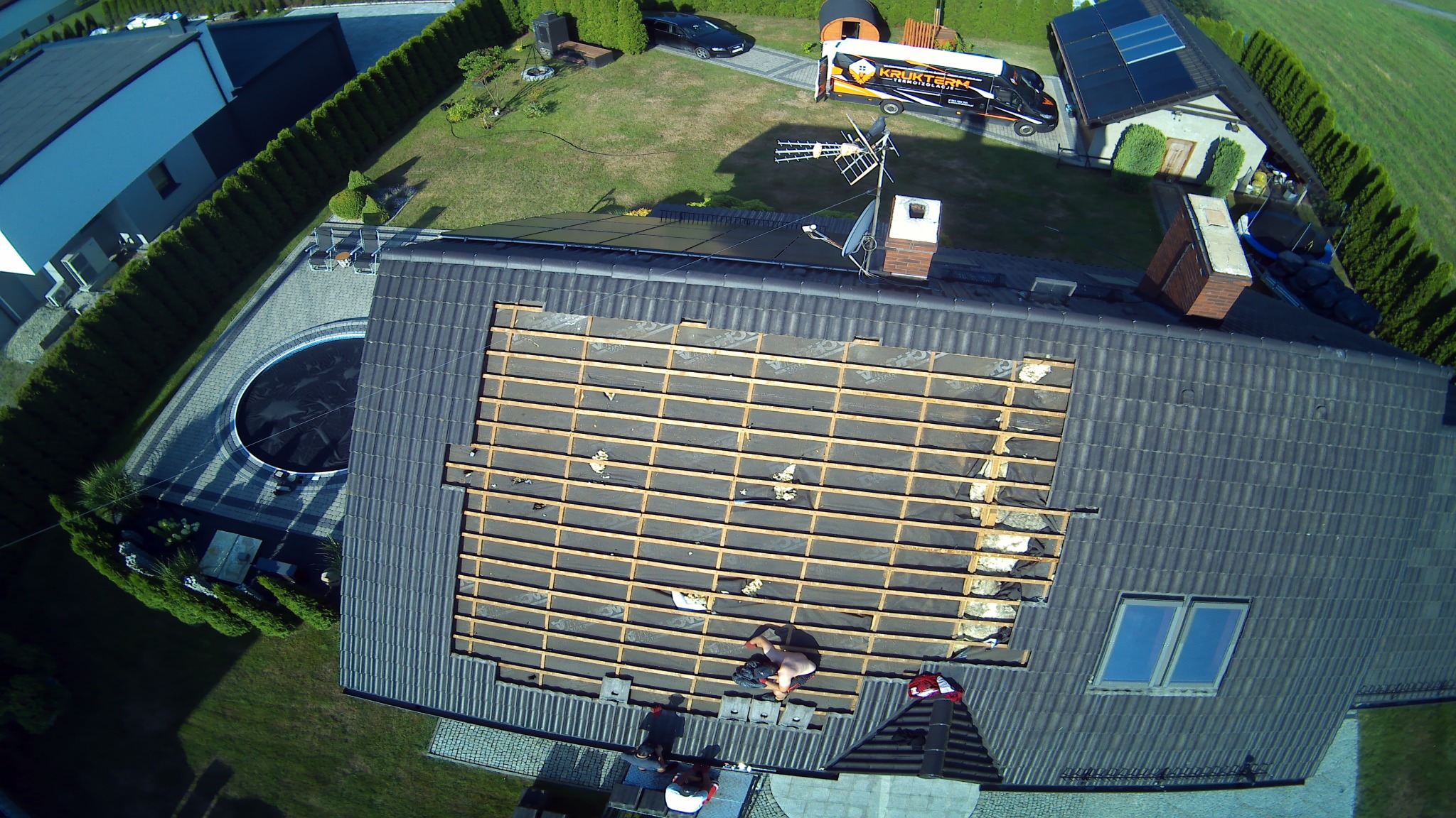 Widok z lotu ptaka na remontowany dach domu z panelami słonecznymi, gdzie widać konstrukcję drewnianą i pracujących robotników, a w tle basen i firmowy samochód.