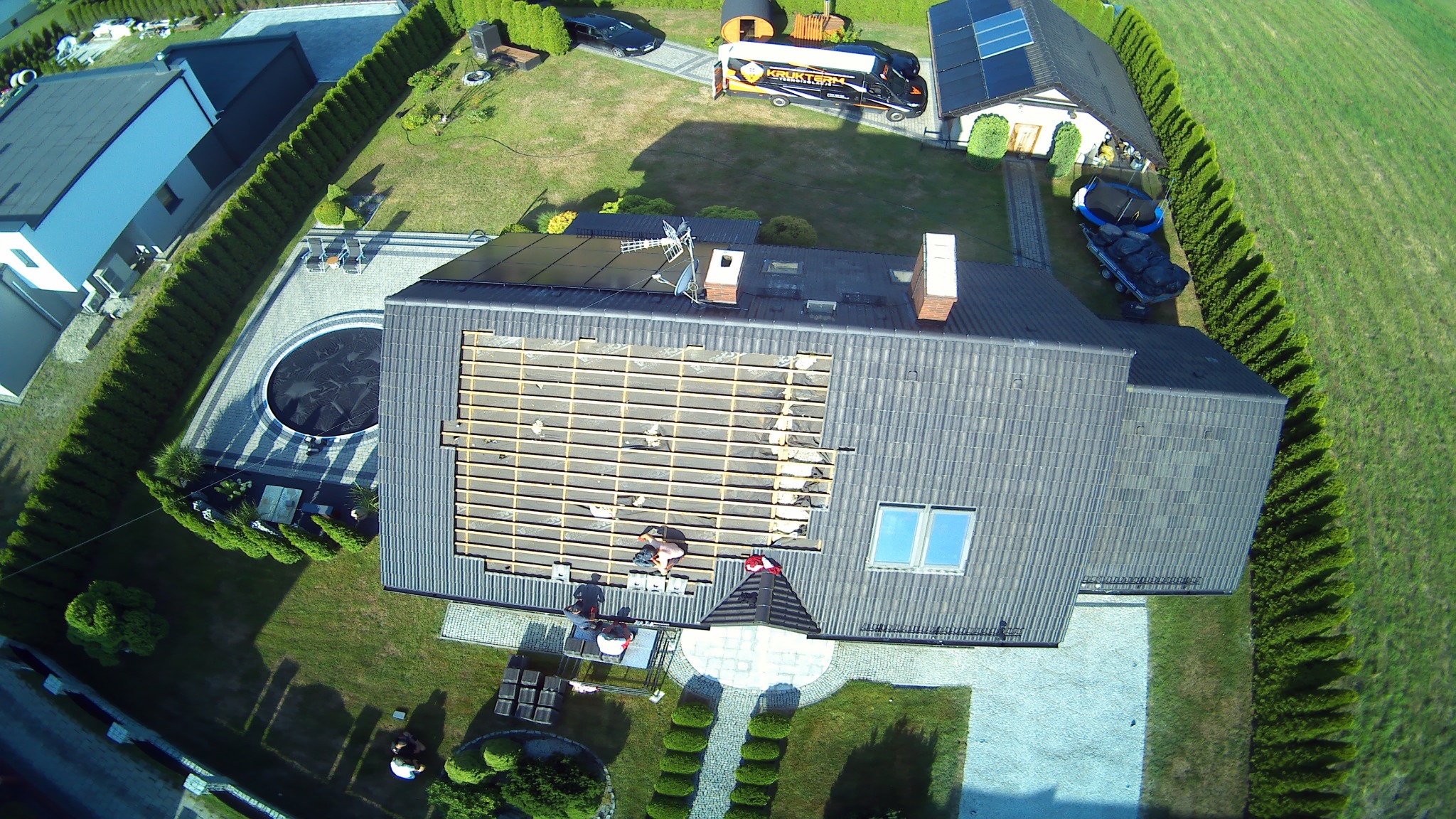 Widok z drona na dom z częściowo zdjętym dachem ukazującym drewnianą konstrukcję, ekipa budowlana na dachu, panele słoneczne, zadbany ogród z basenem i pojazd firmy budowlanej.