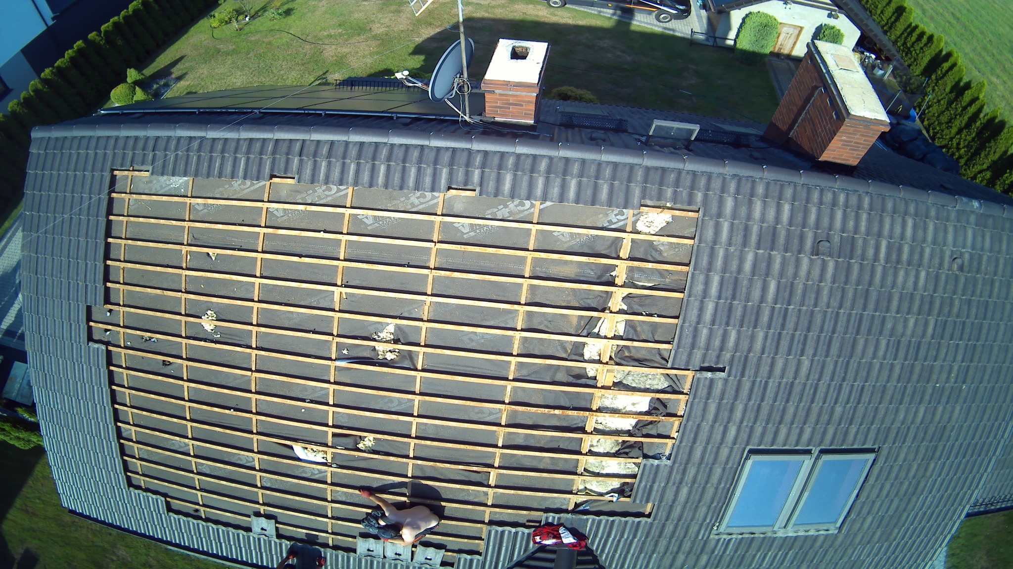 Widok z góry na remontowany dach z widoczną konstrukcją drewnianą, czarną membraną i fragmentami białej izolacji, częściowo pokryty szarą dachówką, z kominem i anteną satelitarną w tle.