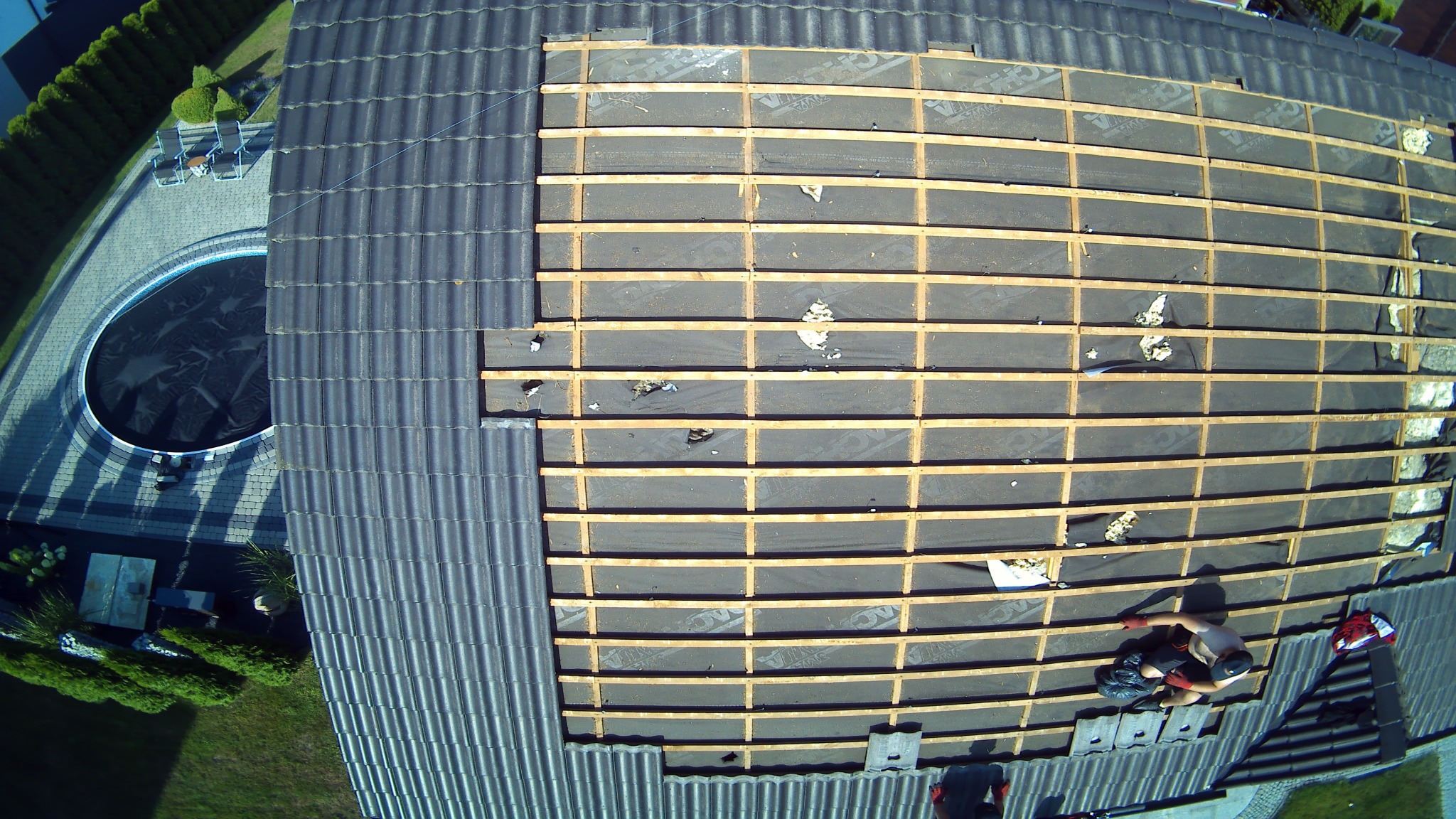 Poddasze z częściowo zdjętym pokryciem, widoczne drewniane łaty i czarna membrana, z fragmentami izolacji. Z góry widoczny pracownik w trakcie pracy, obok basen i zadbany ogród.