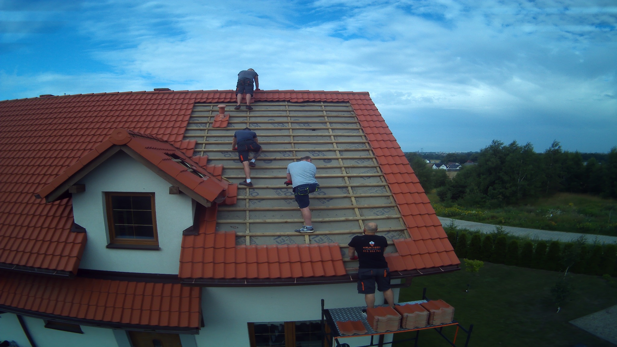 Trzech mężczyzn pracuje na dachu domu z czerwoną dachówką, układając nowe płytki na łatach; widok z góry na budynek i okoliczną zieleń