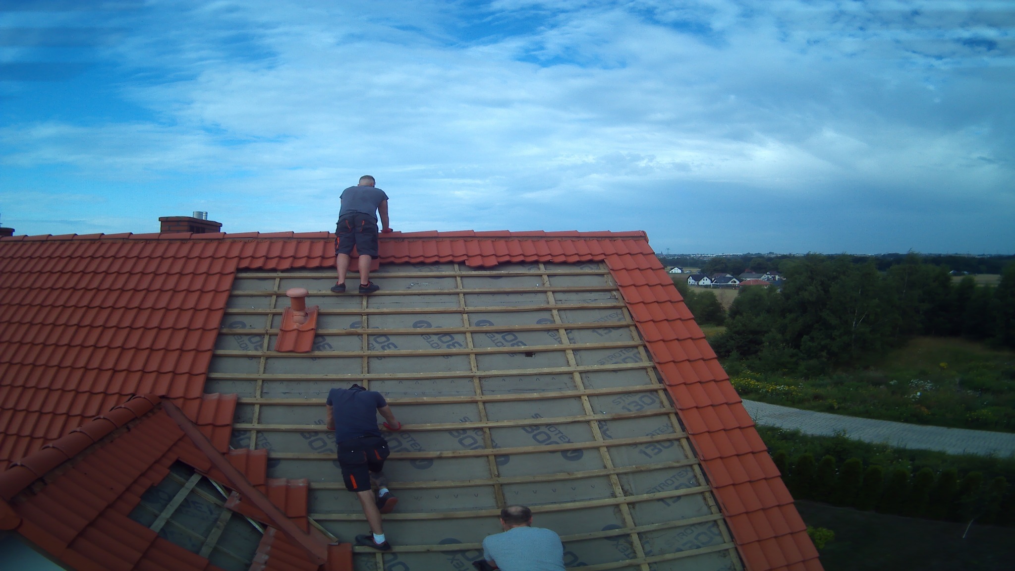 Trzech mężczyzn pracuje na dachu domu z czerwoną dachówką, widoczne drewniane łaty i membrana dachowa, fragment dachu bez dachówek, w tle błękitne niebo z chmurami i zieleń drzew