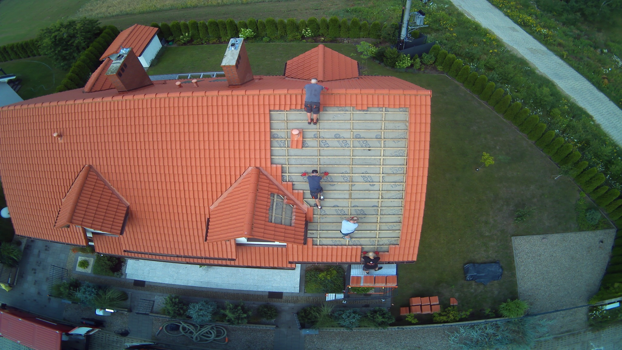 Widok z drona na ekipę montującą izolację pod dachówką na nowym dachu z czerwonej dachówki, widoczne płyty izolacyjne z nadrukiem.