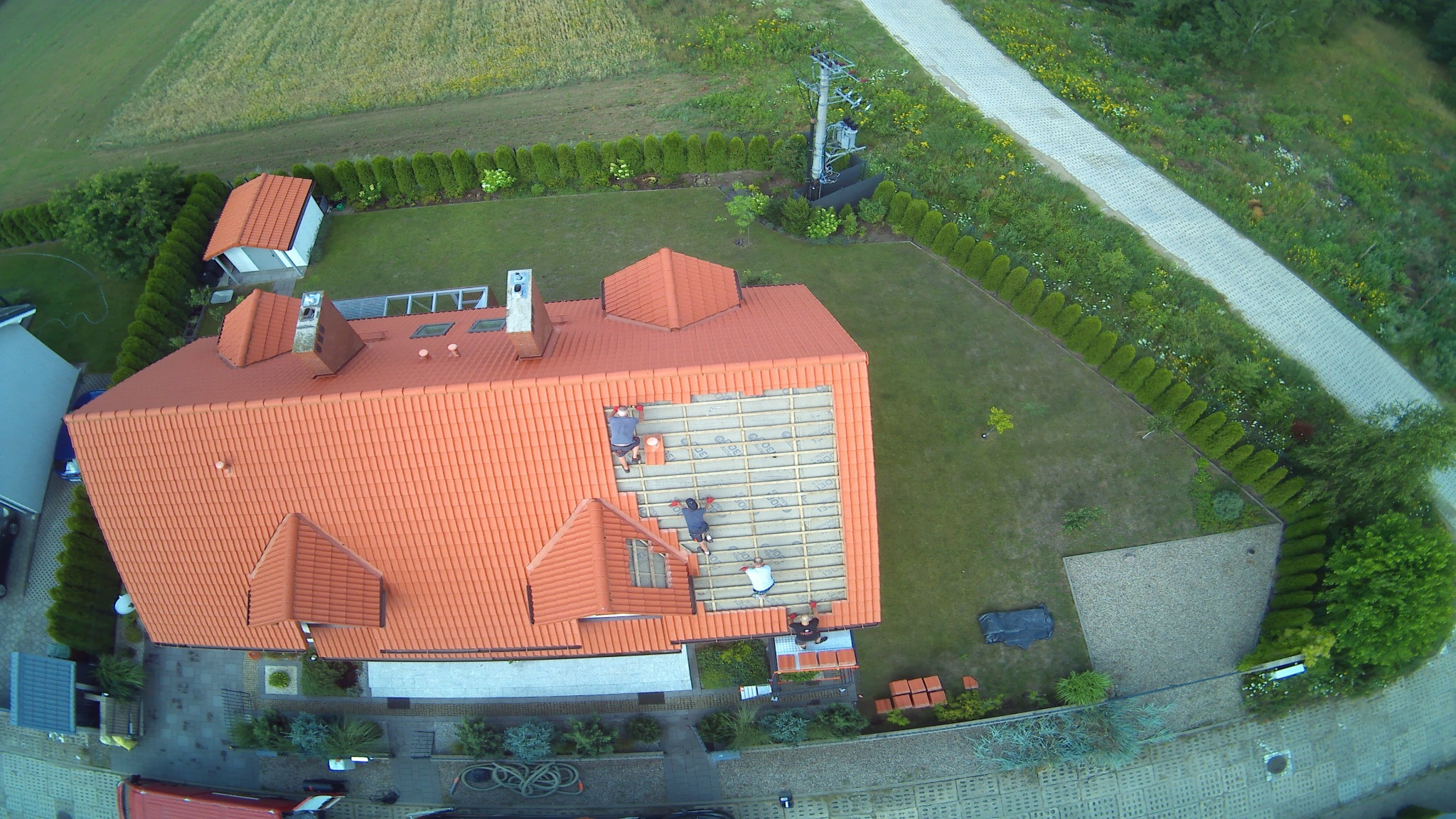 Widok z góry na dom z czerwoną dachówką, gdzie czterech pracowników montuje izolację na fragmencie dachu, na tle zielonego ogrodu i pola.
