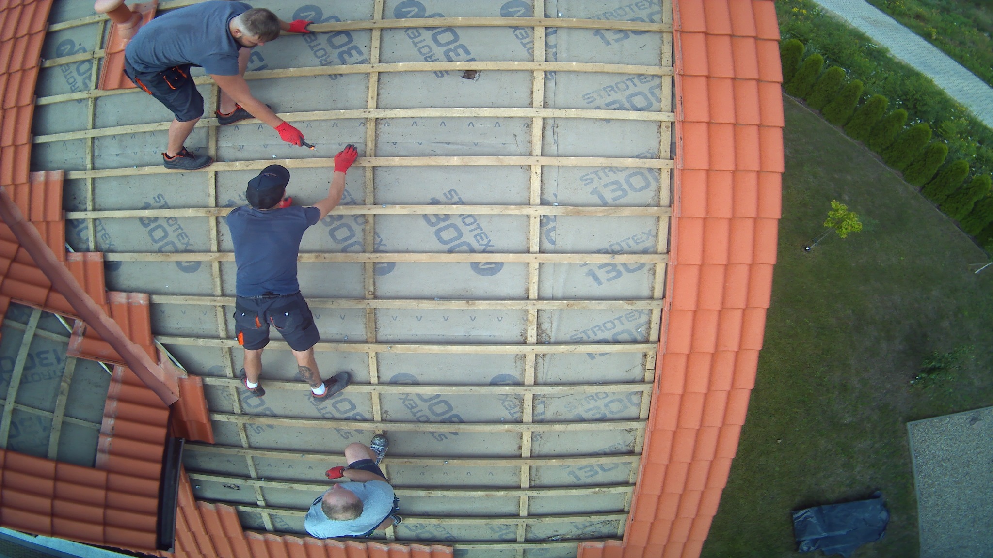 Montaż izolacji na poddaszu: trzech mężczyzn w czerwonych rękawicach mocuje folię Strotex 1300 do drewnianej konstrukcji dachu, widok z góry na budynek z pomarańczową dachówką i zielonym trawnikiem.