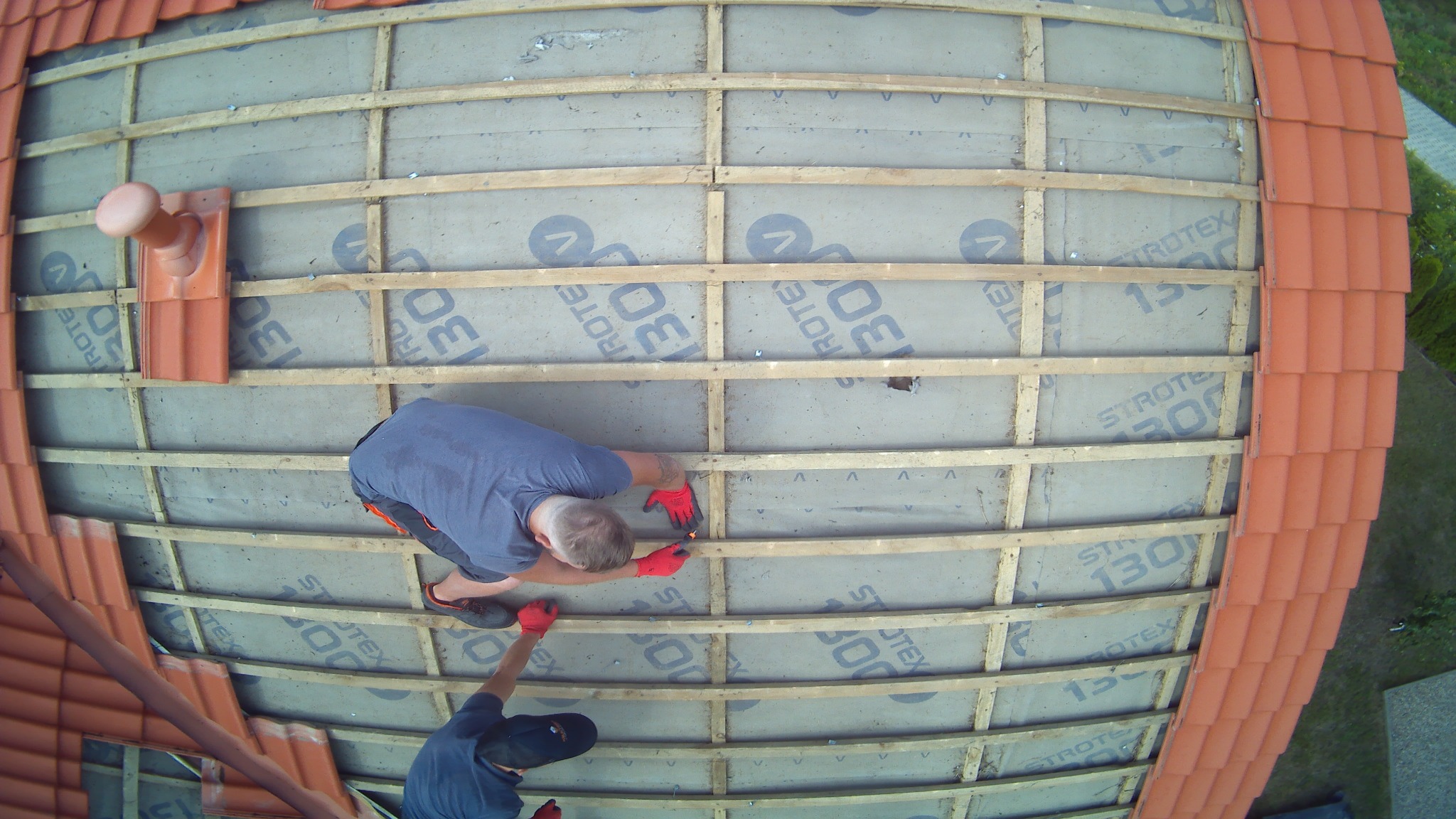 Widok z góry na dwóch robotników w czerwonych rękawicach montujących drewnianą konstrukcję dachu z membraną Strotex 130, z częściowo ułożoną czerwoną dachówką.