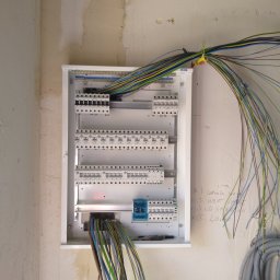 DoSo Elektryk Roman Moskaliuk - Podłączenie przewodów do rozdzielnicy elektrycznej na surowej ścianie, widoczne zabezpieczenia EATON i notatki ołówkiem na ścianie.
