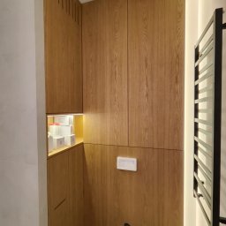 DoSo Elektryk Roman Moskaliuk - Nowoczesna łazienka z zabudową z drewnianych paneli, oświetleniem LED i grzejnikiem drabinkowym w czarnym kolorze. Widoczny fragment stelaża podtynkowego WC i otwarta półka z kosmetykami.