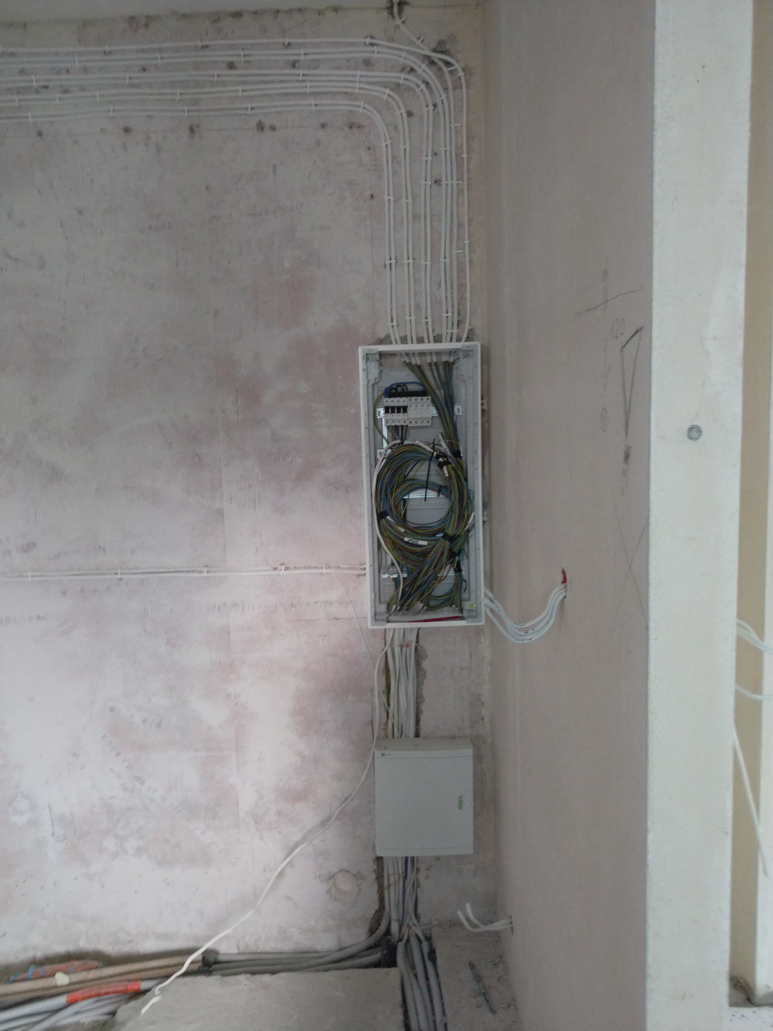 Rozdzielnica elektryczna z podłączonymi przewodami w surowym wnętrzu, widoczne kable prowadzone po ścianie w białych rurkach, dolna skrzynka zasilająca.
