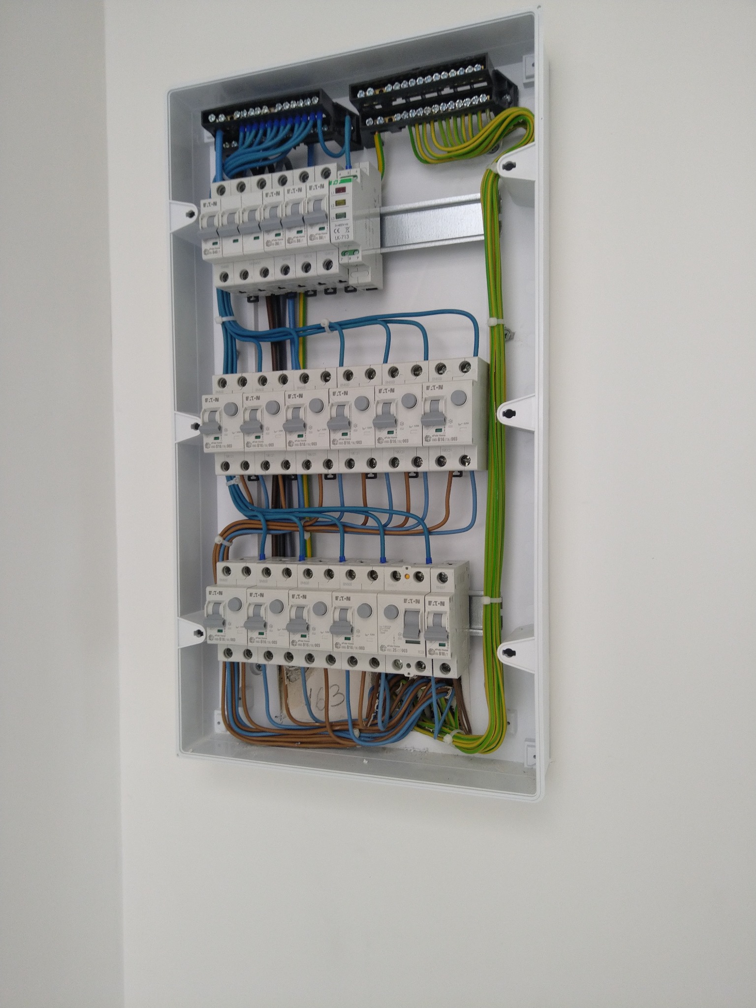 Rozdzielnica elektryczna z bezpiecznikami i okablowaniem, zamontowana na białej ścianie w Warszawie. Widoczne niebieskie, brązowe i zielono-żółte przewody oraz białe wyłączniki firmy Eaton.