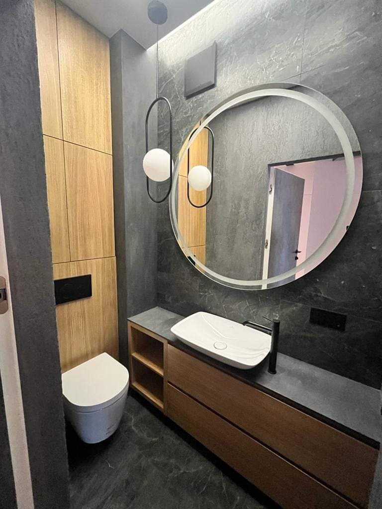 Nowoczesna łazienka w odcieniach szarości i drewna, z okrągłym podświetlanym lustrem, wiszącą lampą z dwoma kulami, białą umywalką nablatową i zabudowaną toaletą.