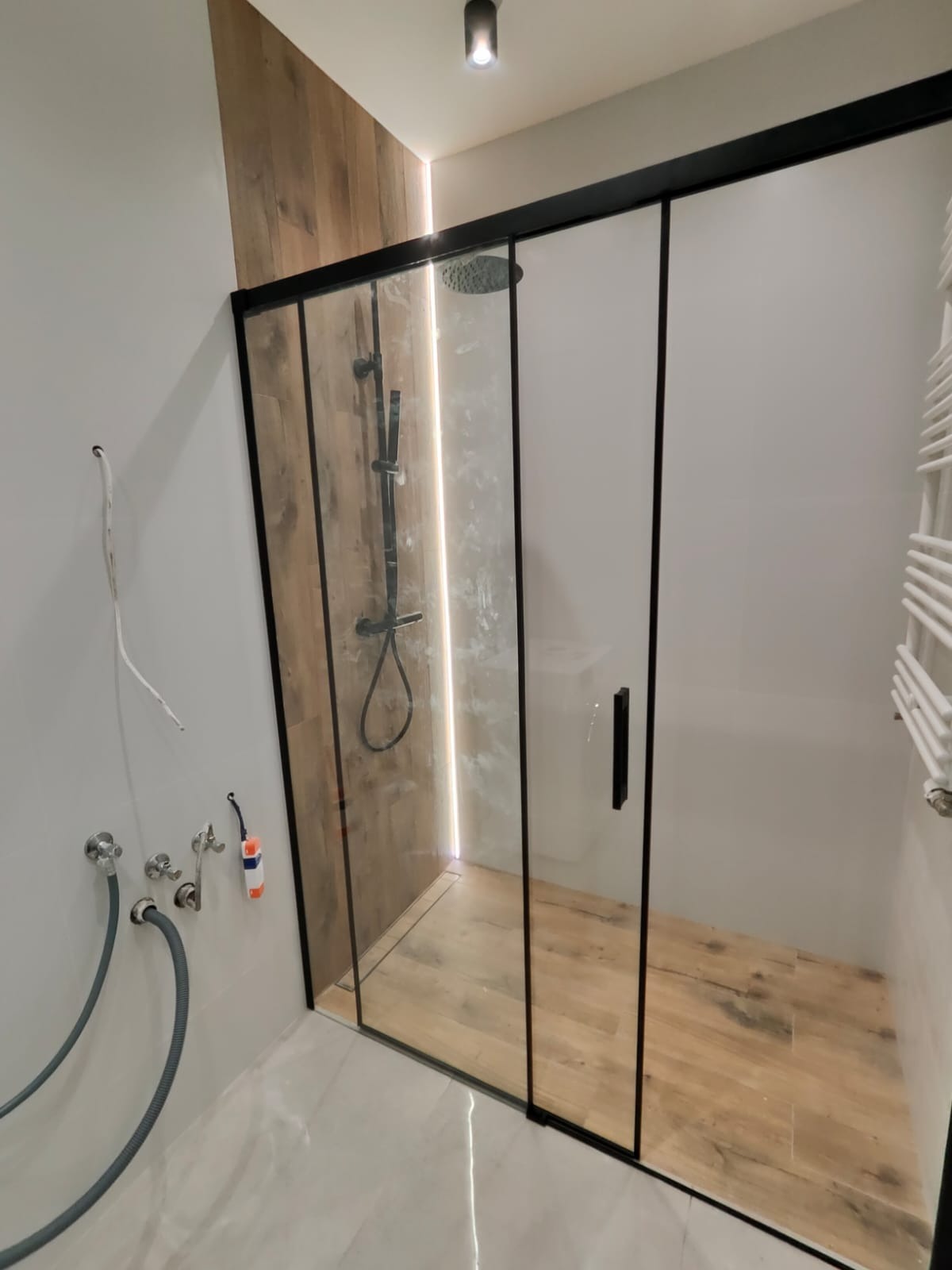 Nowoczesna kabina prysznicowa z czarnymi profilami, drewnopodobną ścianą i podłogą, widoczne przyłącza wodne i odpływ.