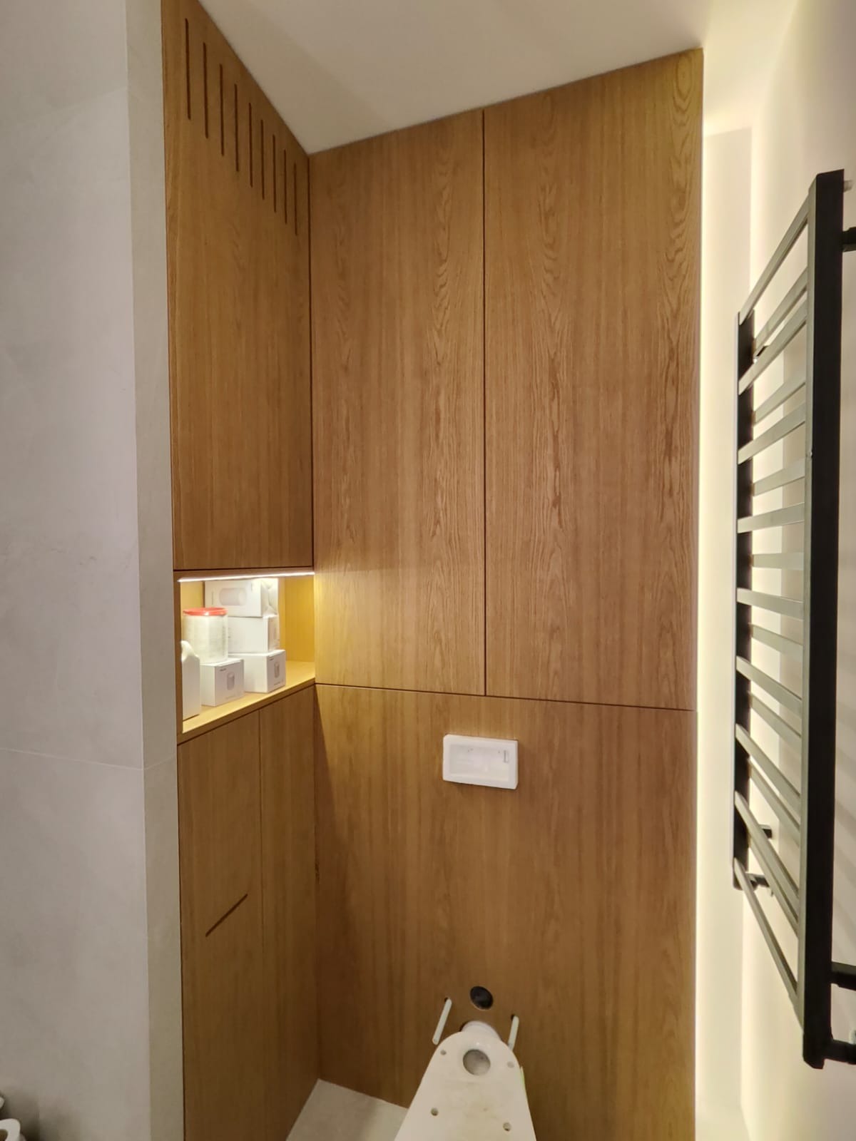 Nowoczesna łazienka z zabudową z drewnianych paneli, oświetleniem LED i grzejnikiem drabinkowym w czarnym kolorze. Widoczny fragment stelaża podtynkowego WC i otwarta półka z kosmetykami.