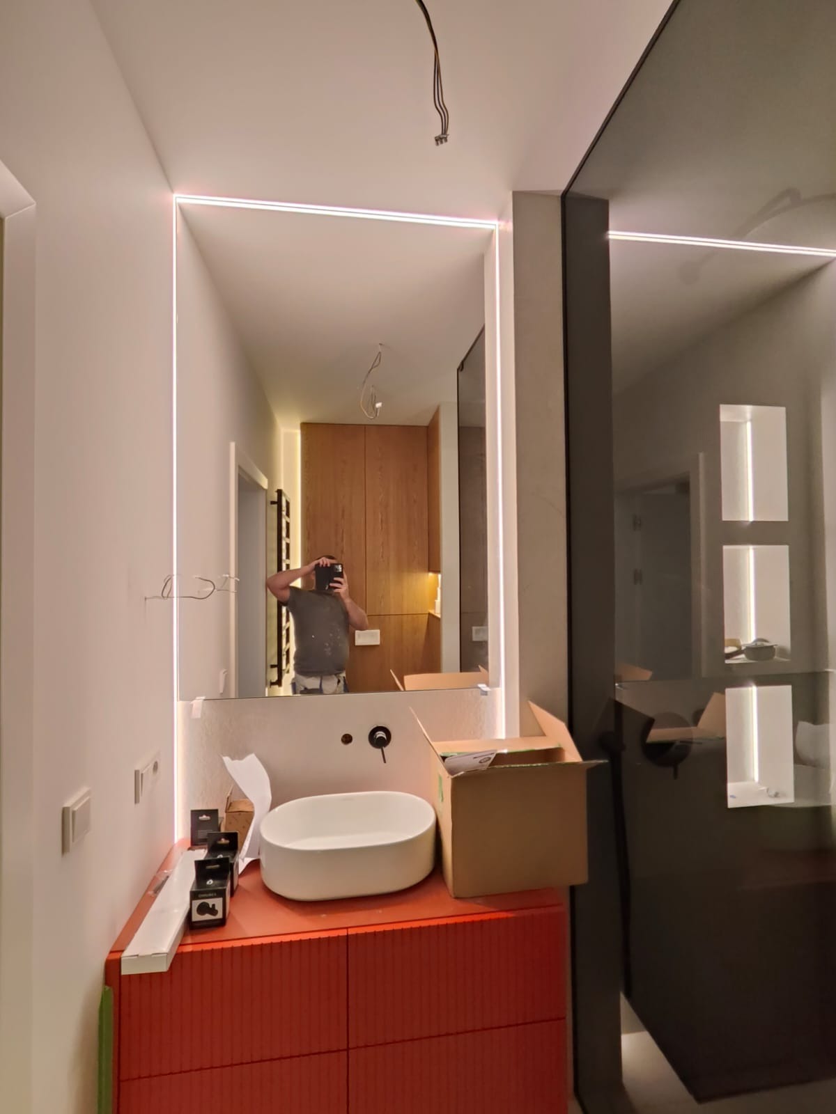 Łazienka w trakcie remontu z podświetlonym lustrem LED, widoczne kartony i akcesoria montażowe na pomarańczowej szafce pod umywalką, instalacja elektryczna w suficie.