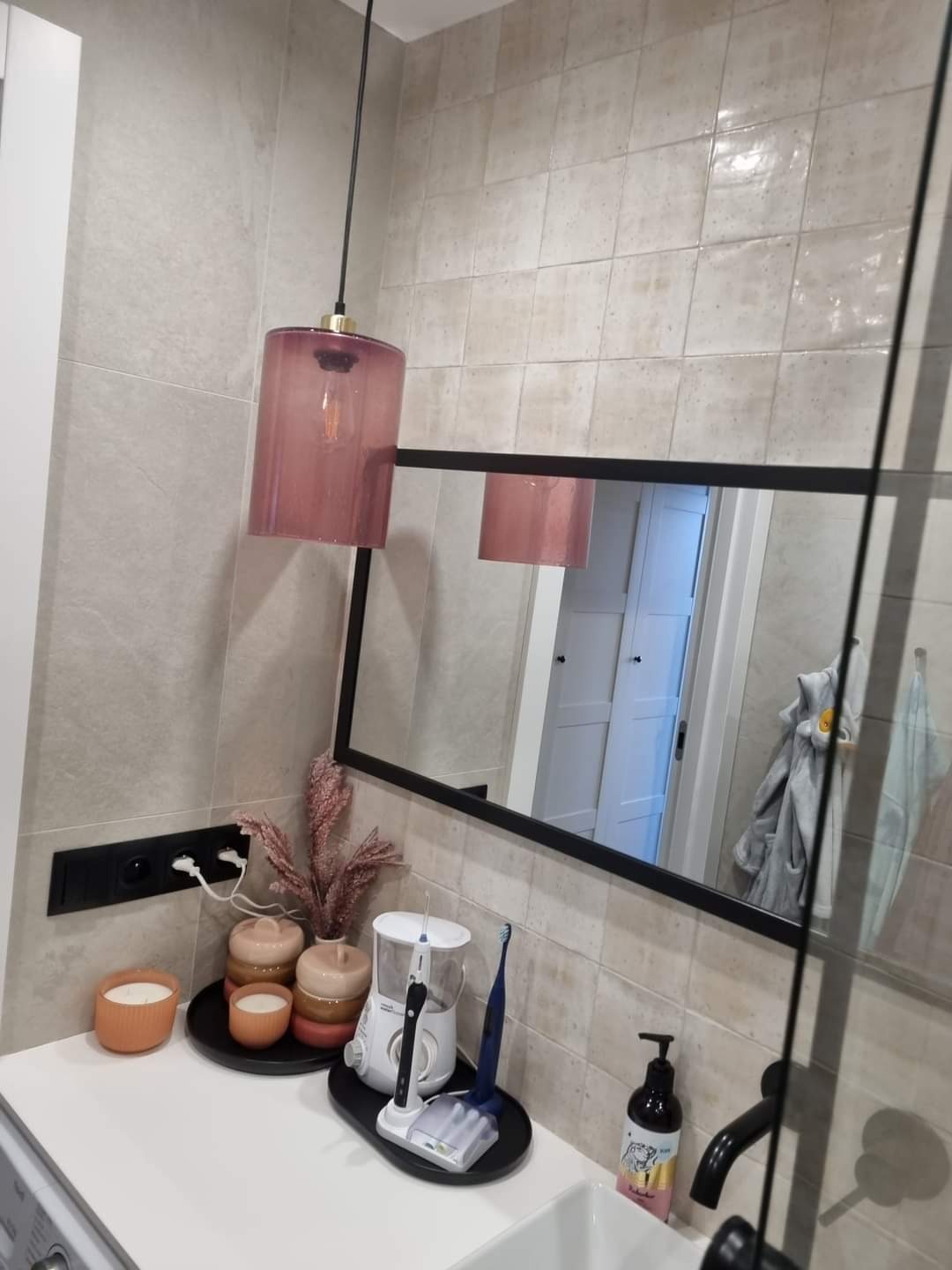 Nowoczesna łazienka z wiszącą lampą w kolorze pudrowego różu, czarną oprawą lustra i elementami dekoracyjnymi na blacie umywalki.