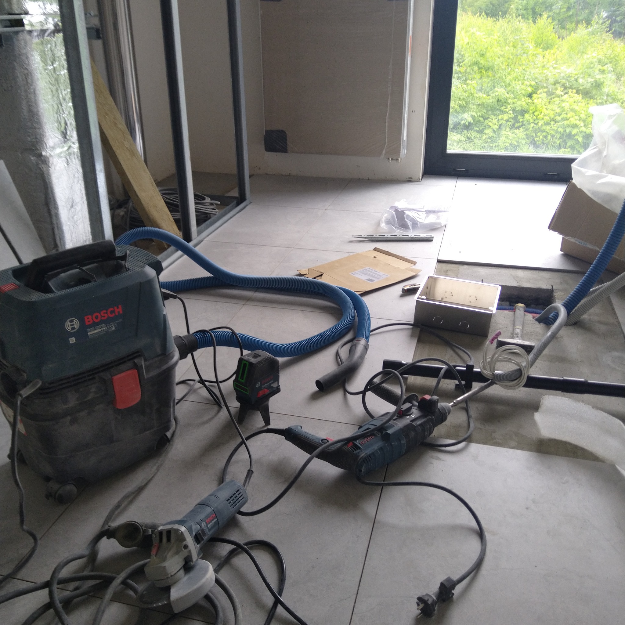 Bosch odkurzacz przemysłowy, szlifierka kątowa i wiertarka udarowa leżące na podłodze w trakcie prac wykończeniowych, widoczna skrzynka elektryczna i laser krzyżowy.