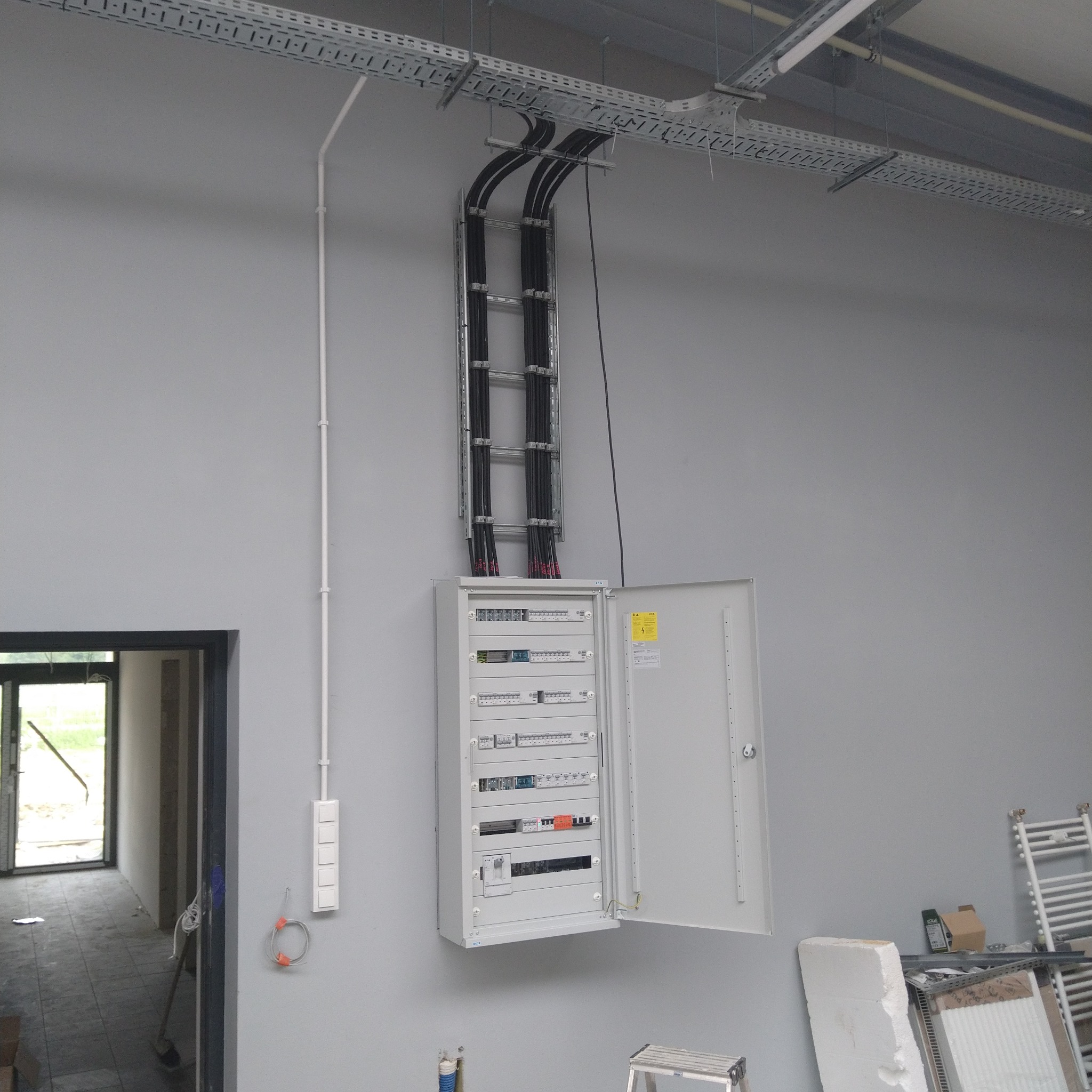 Szara ściana z rozdzielnicą elektryczną z otwartymi drzwiami, widoczne kable w korycie i peszlu, w tle surowy stan wykończeniowy pomieszczenia.