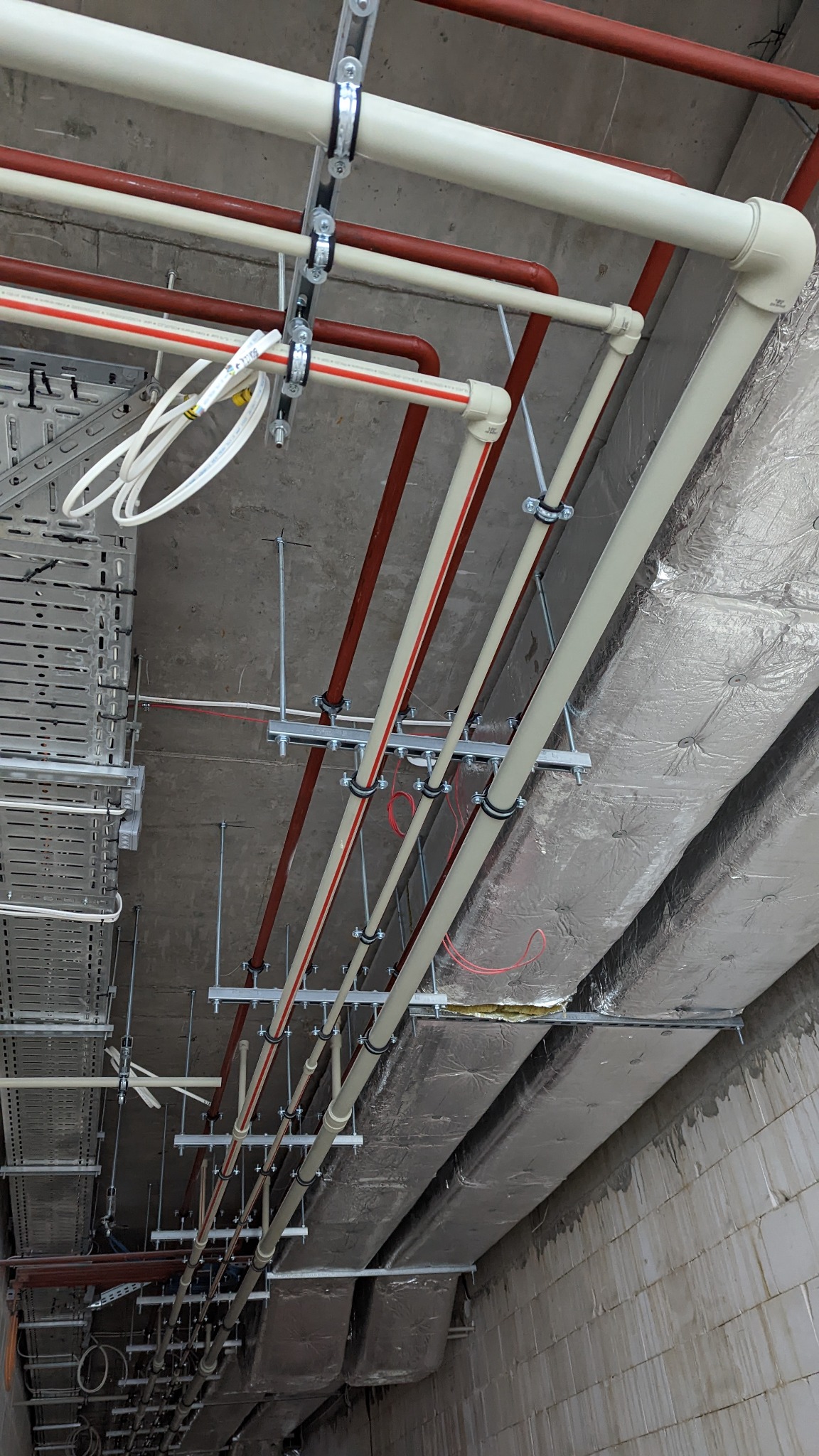 Kompleksowy montaż instalacji hydraulicznych i wentylacyjnych pod sufitem w Warszawie, widoczne rury w kolorze beżowym i czerwonym, podwieszane na metalowych wspornikach, oraz izolowane kanały...