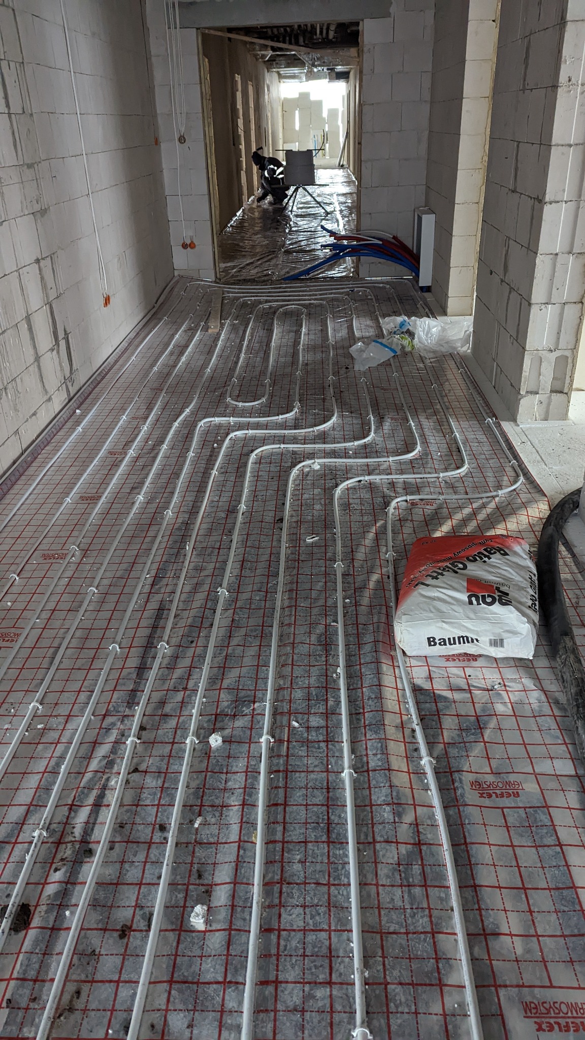 Rozłożone rury ogrzewania podłogowego na folii z siatką w surowym wnętrzu, widoczny worek z zaprawą Baumit, w tle pracownik w kombinezonie.