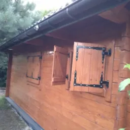 Drewniana altana ogrodowa z otwartymi okiennicami z czarnymi okuciami, widok z zewnątrz na tle zieleni.