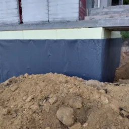 Fundament budynku zabezpieczony izolacją przeciwwilgociową i termiczną, z widoczną folią kubełkową i stertą ziemi.