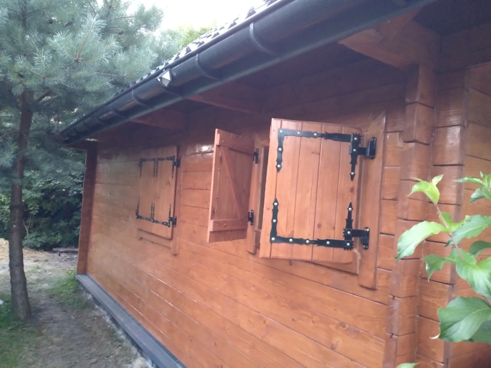 Drewniana altana ogrodowa z otwartymi okiennicami z czarnymi okuciami, widok z zewnątrz na tle zieleni.