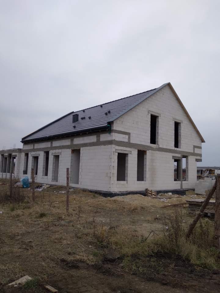 Surowy stan budynku jednorodzinnego z szarym dachem z dachówki, widoczne niezabezpieczone otwory okienne i drzwiowe, ściany z białego pustaka, teren wokół niezagospodarowany.