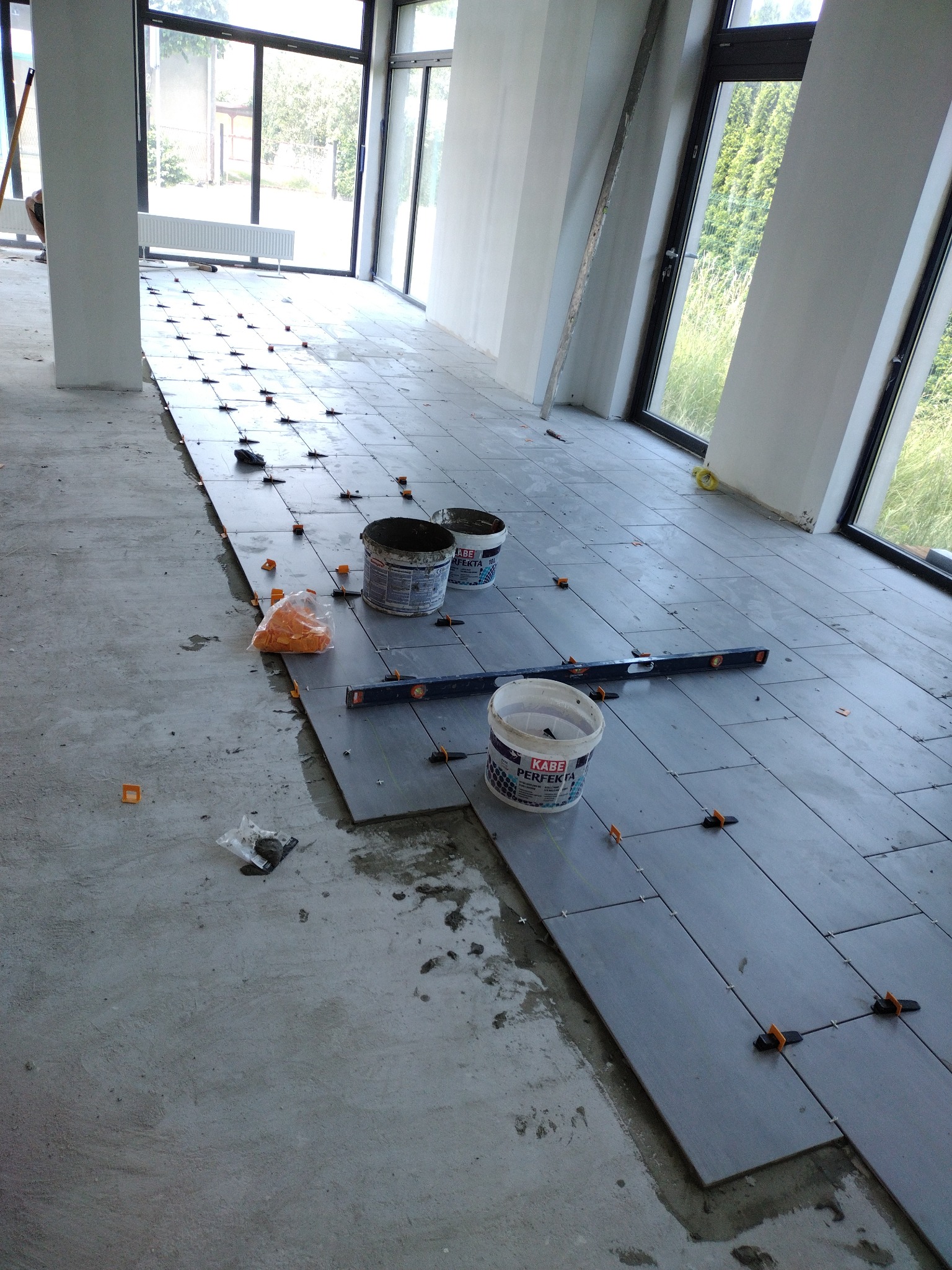 Wnętrze w trakcie układania szarych płytek podłogowych z użyciem poziomicy i plastikowych krzyżyków dystansowych; widoczne otwarte wiadra z klejem do płytek oraz woreczek z krzyżykami.