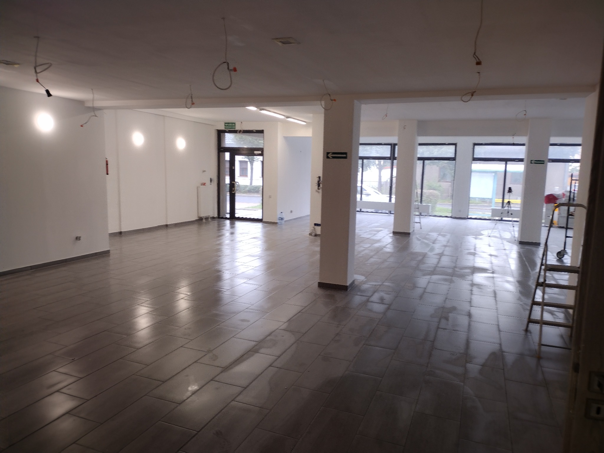 Przestronne, jasne wnętrze po remoncie z ułożoną szarą podłogą z płytek imitujących panele, widoczne niedokończone instalacje elektryczne pod sufitem, białe ściany i kolumny oraz drabina malarska...