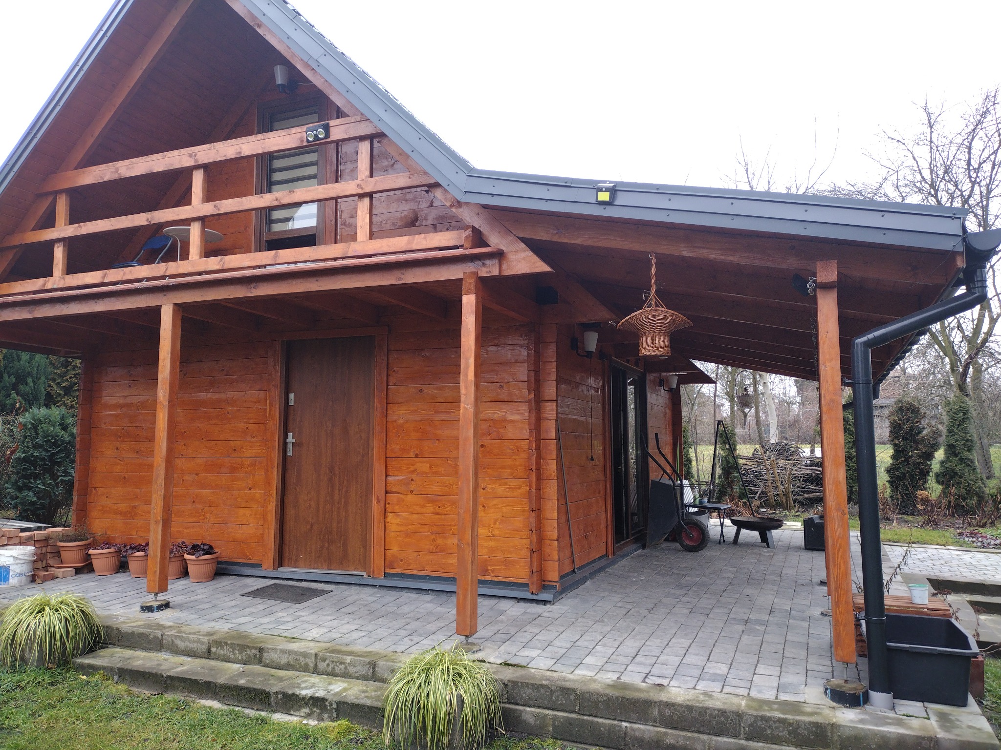 Drewniany domek letniskowy z balkonem, widok z zewnątrz, zadaszony taras wyłożony kostką brukową, rynna odprowadzająca wodę, rośliny w donicach i elementy wyposażenia ogrodu w tle.