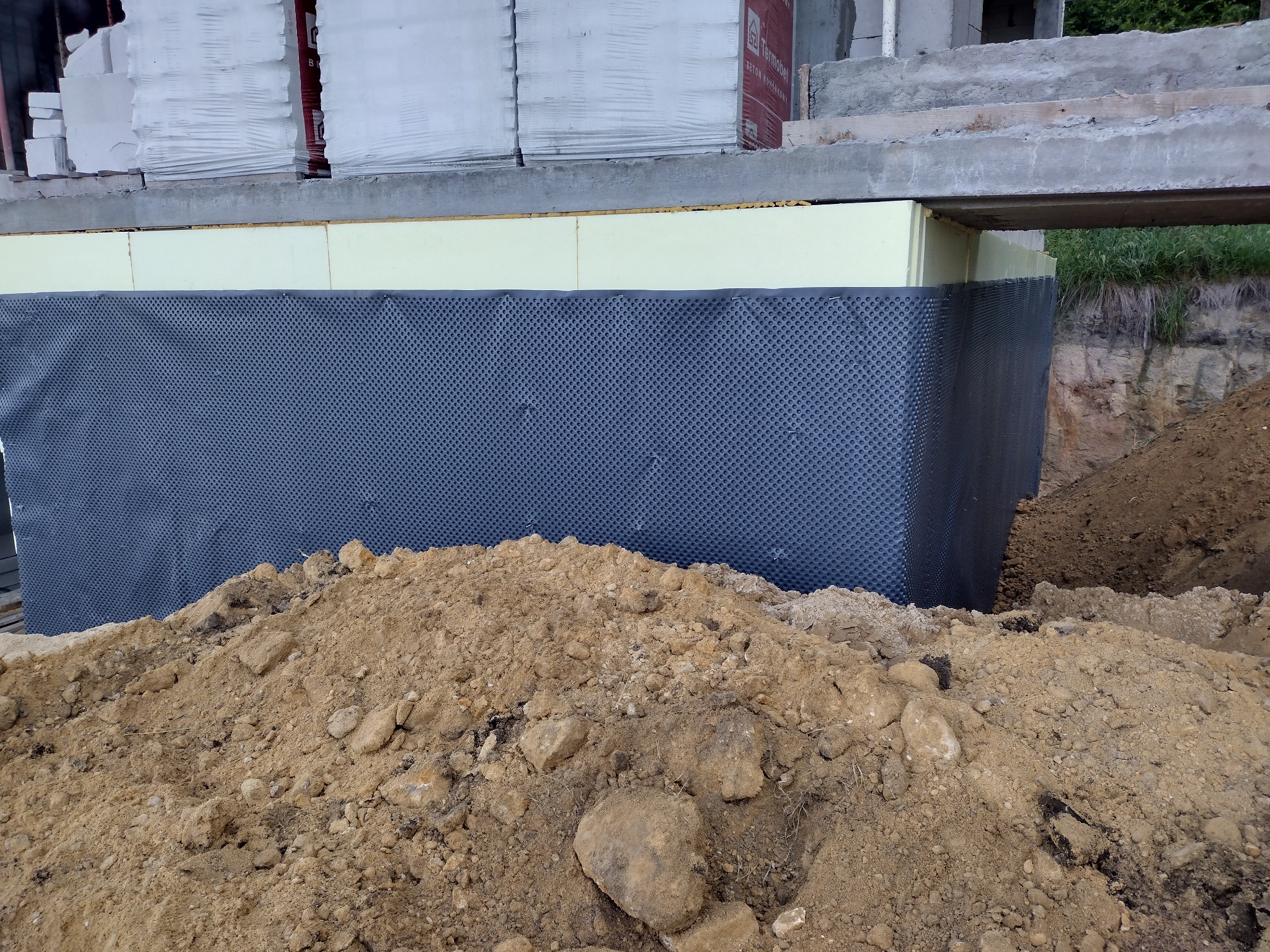 Fundament budynku zabezpieczony izolacją przeciwwilgociową i termiczną, z widoczną folią kubełkową i stertą ziemi.