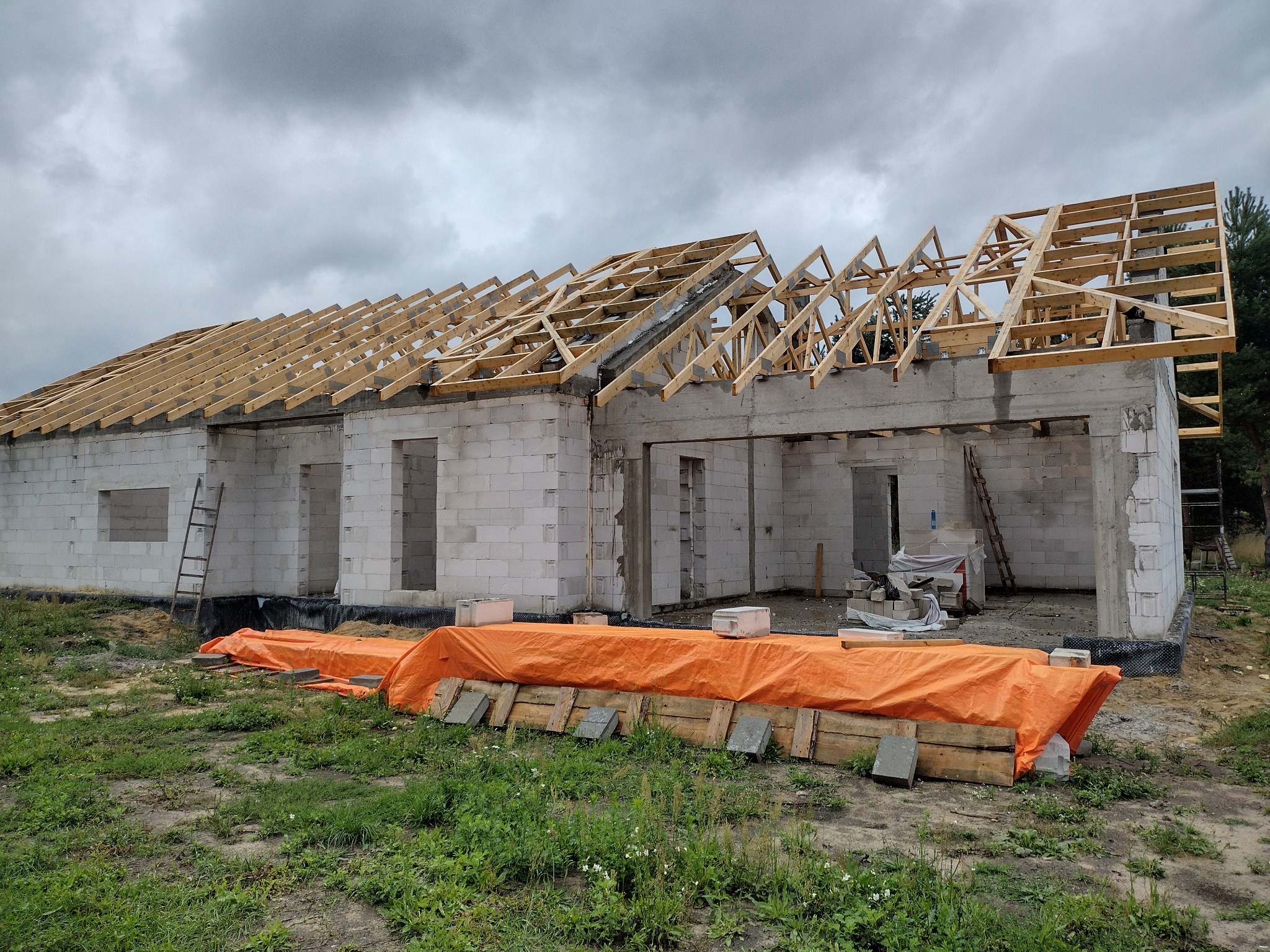 Surowy stan budynku jednorodzinnego z drewnianą konstrukcją dachu w trakcie montażu, widoczne bloczki betonowe ścian i pomarańczowa plandeka zabezpieczająca teren budowy, drabina oparta o ścianę.