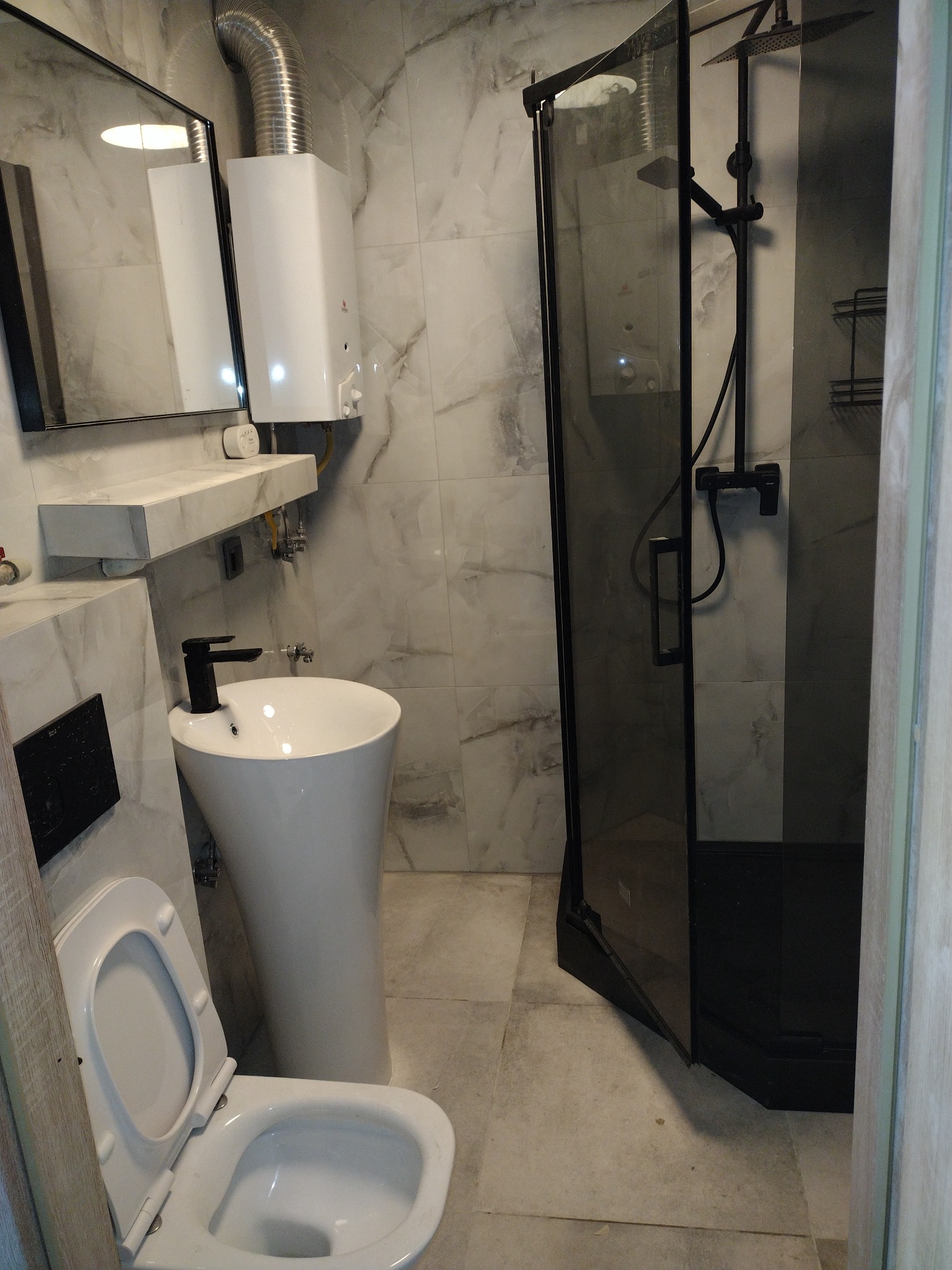 Nowoczesna łazienka z kabiną prysznicową typu walk-in z czarnymi elementami, umywalką wolnostojącą na tle marmurowych płytek i otwartą toaletą.