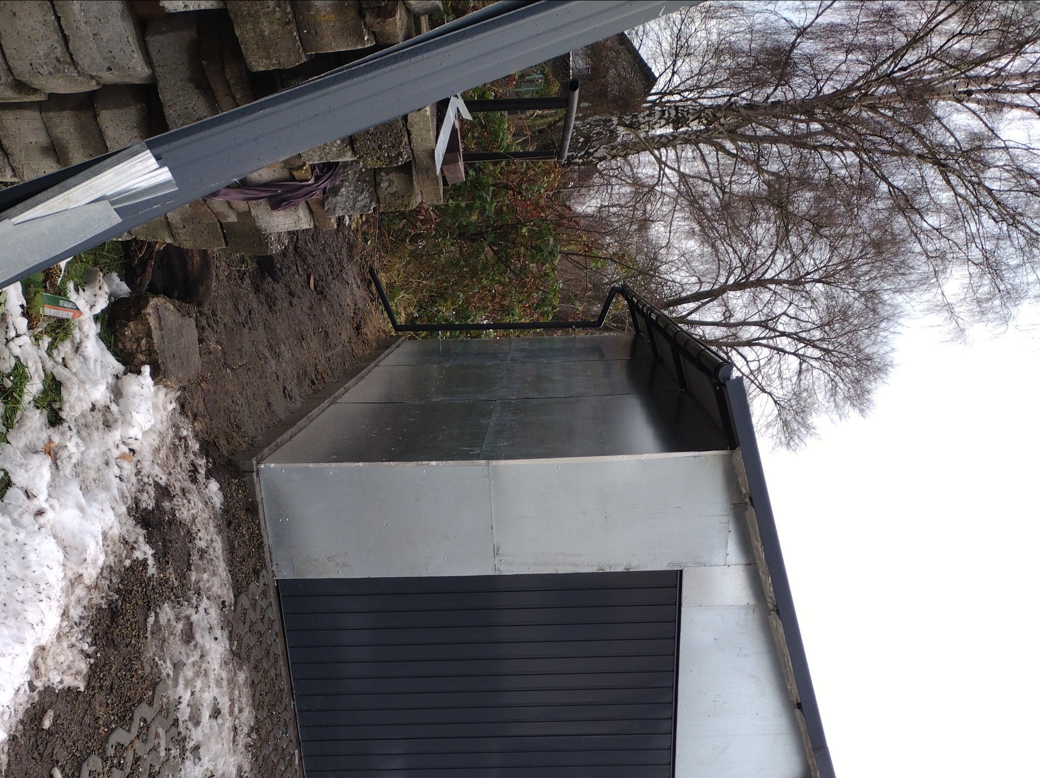 Metalowa konstrukcja dachu z czarną bramą garażową, widoczna z dołu, z fragmentami śniegu na ziemi i drzewem w tle.