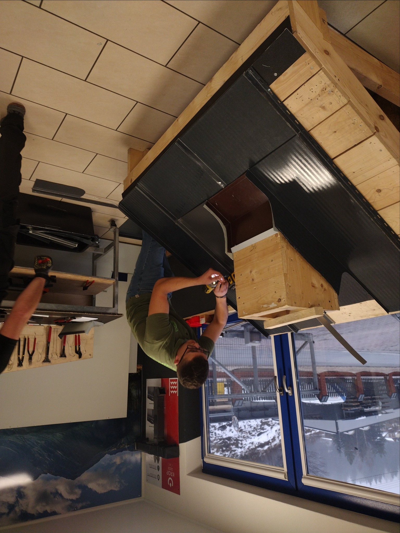 Monteraż blachy na dachu z drewnianą konstrukcją komina wentylacyjnego, widok z wnętrza budynku przez okno na zaśnieżony krajobraz.